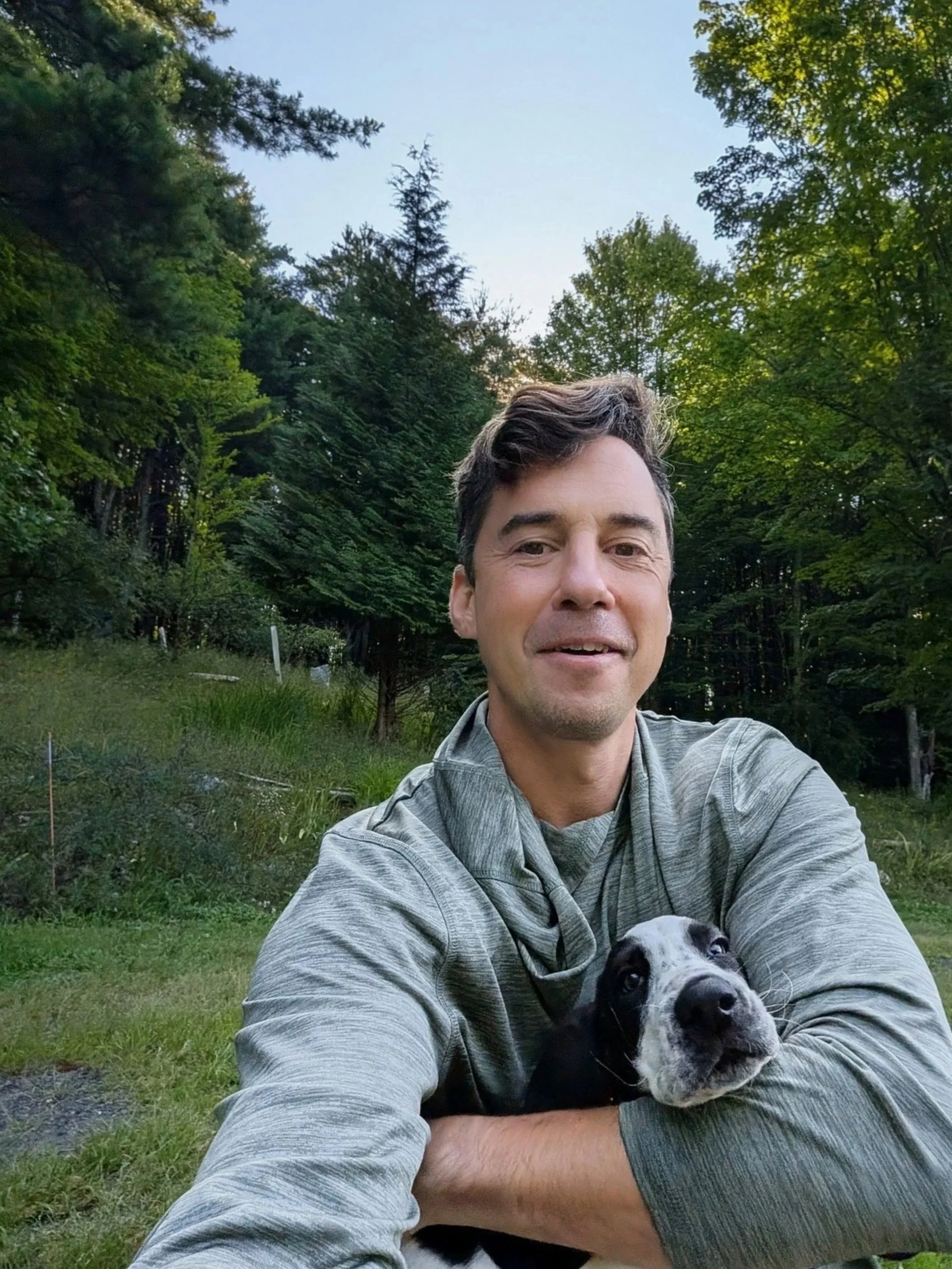 A man taking a selfie outdoors in a green, forested area, holding a black and white puppy.