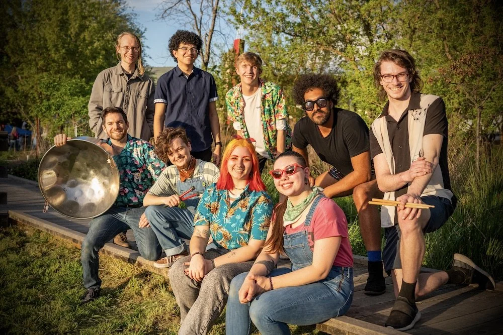 Members of Maraval Road Steel Band sitting outside amongst trees, smiling, with Reed Bentley holding a pan.