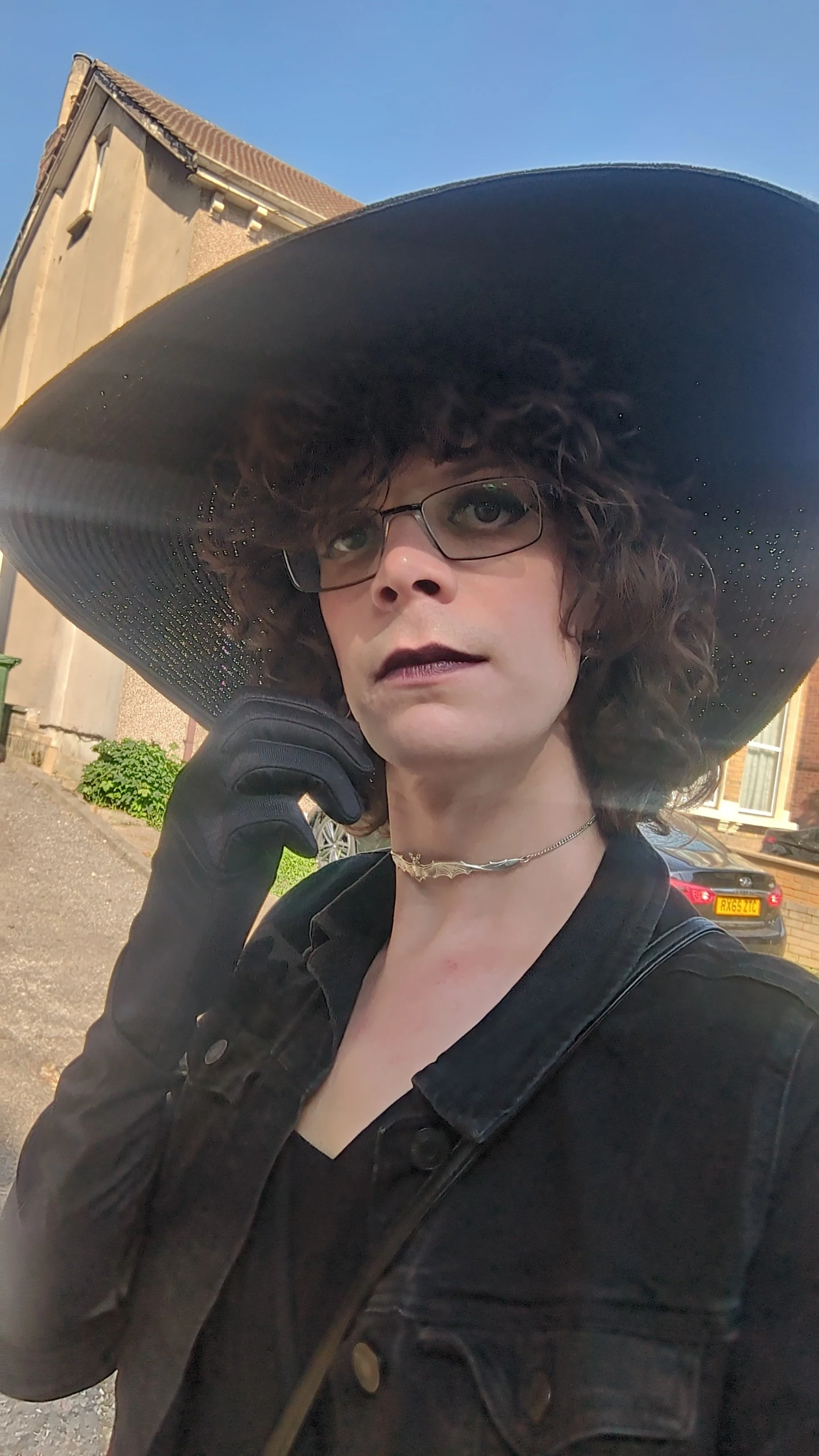 Woman with shoulder-length curly hair, dressed in all black with long, black gloves and a black, wide-brimmed sunhat.