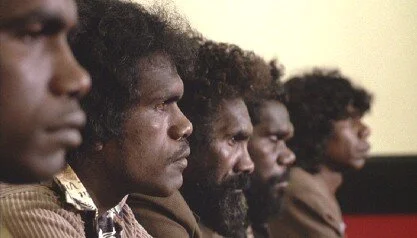 Indigenous men stand in a line looking straight ahead.