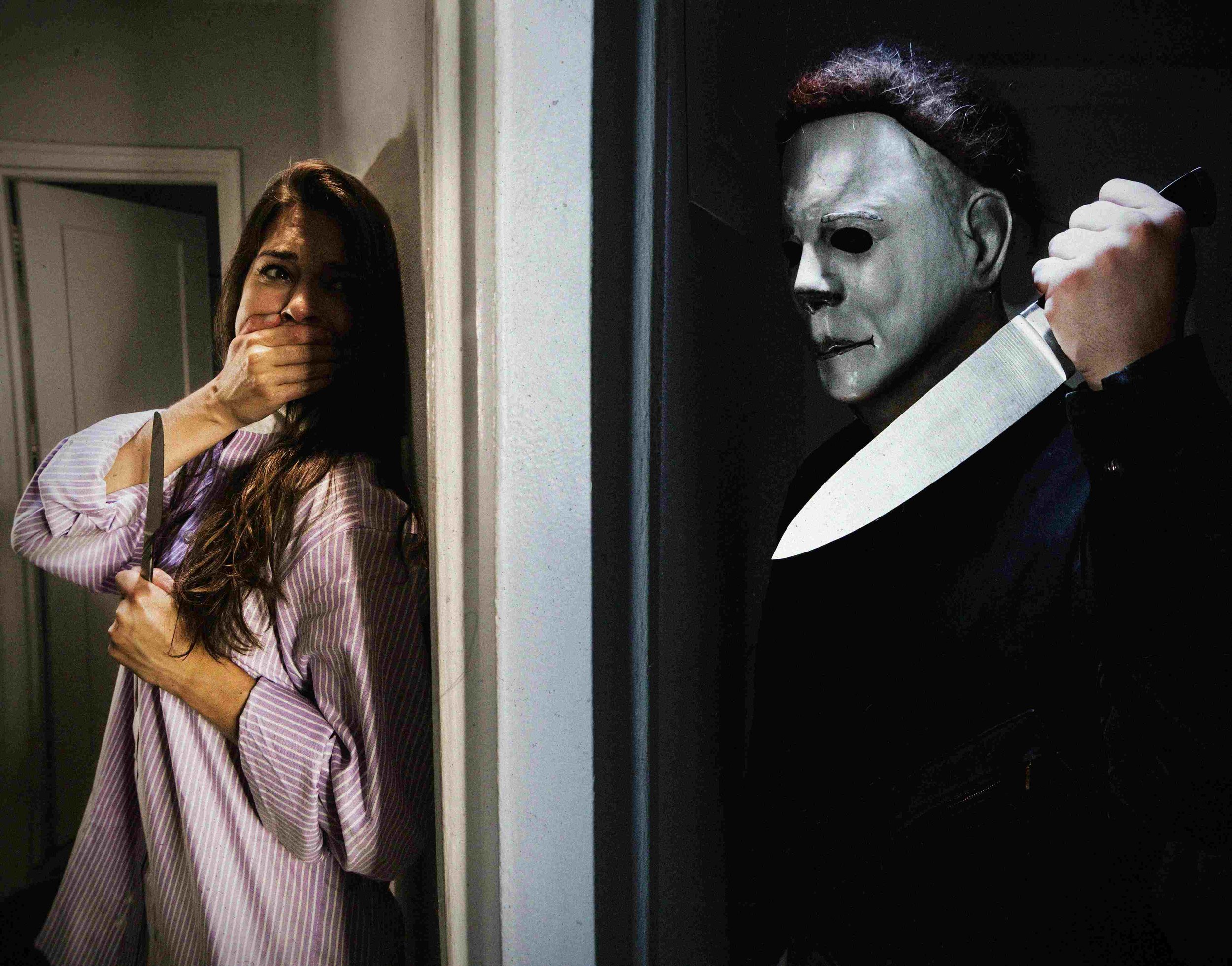 Michael Myers stands in the shadows, knife raised, while his victim stands on the other side of the wall, hand over her mouth.