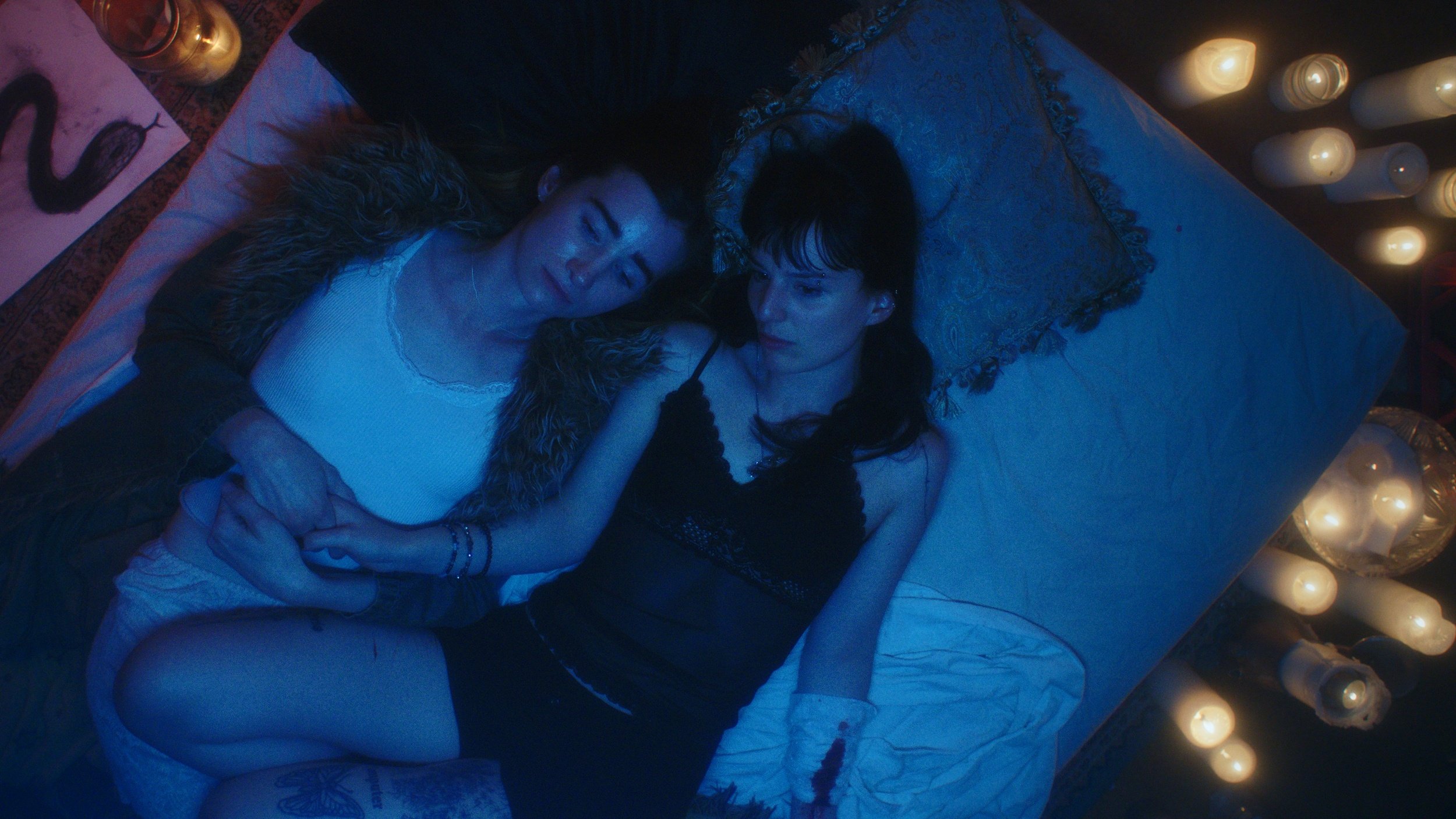 Two women lay on a bed, holding hands. The room is covered in blue light.