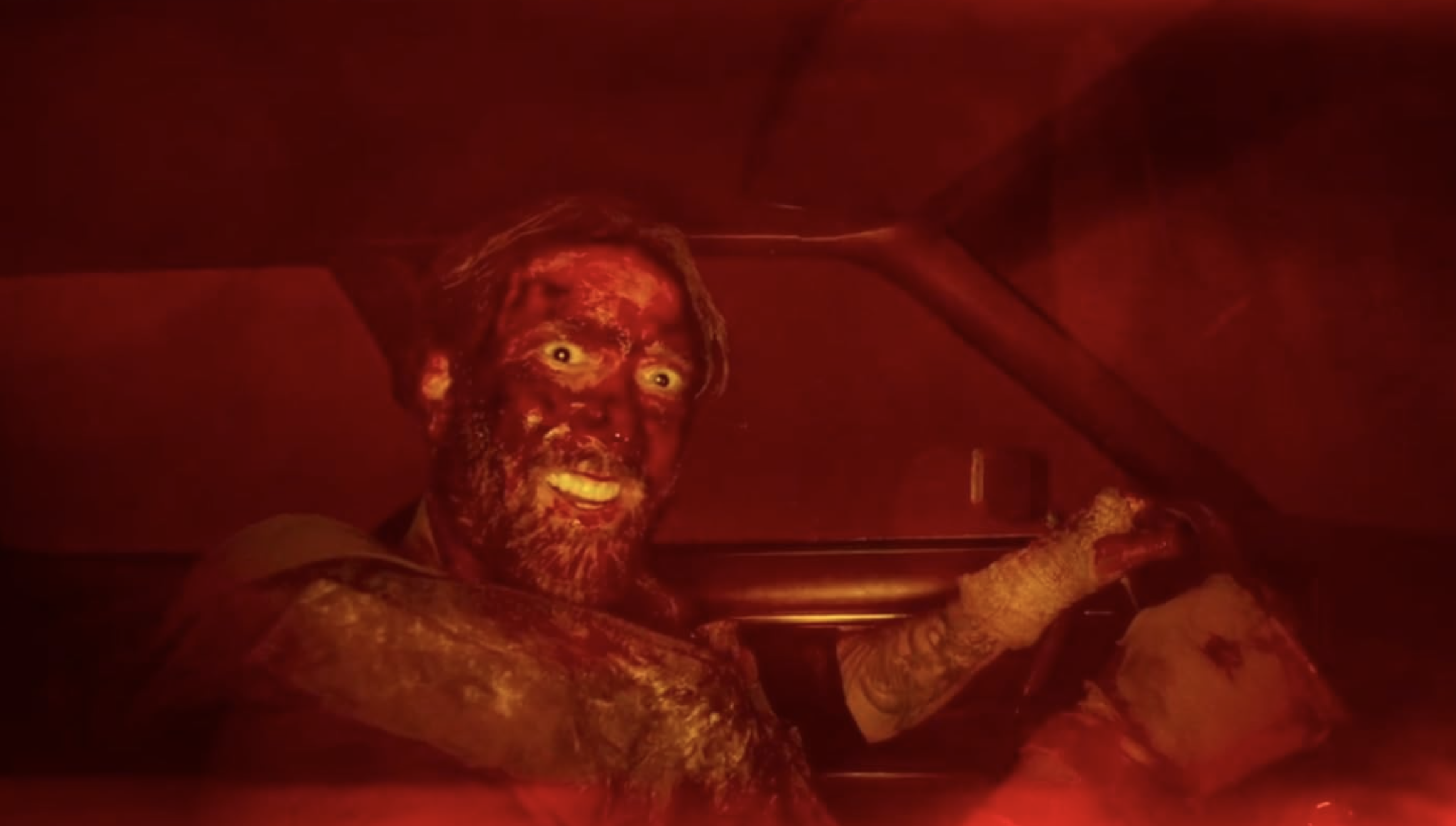 A man with blood covering his face smiles at the camera while he is driving a car.
