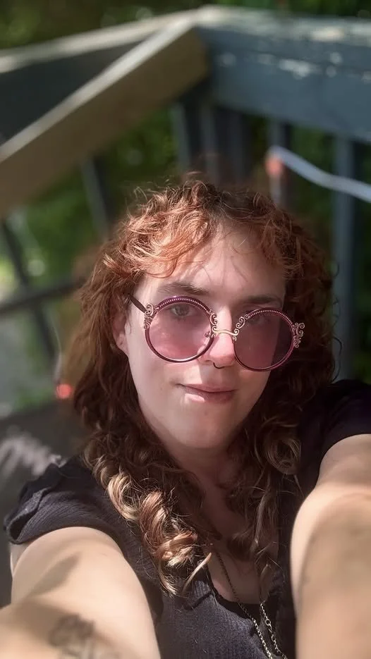 Selfie of a person with long, curly red hair and round rose-colored glasses wearing a black blouse.
