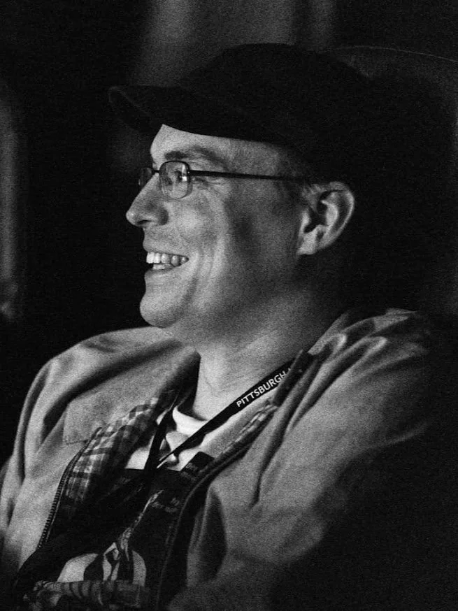 A young man with glasses smiling while holding a phone, sitting at a table in a brick-walled restaurant with wooden ceiling beams and hanging lights.