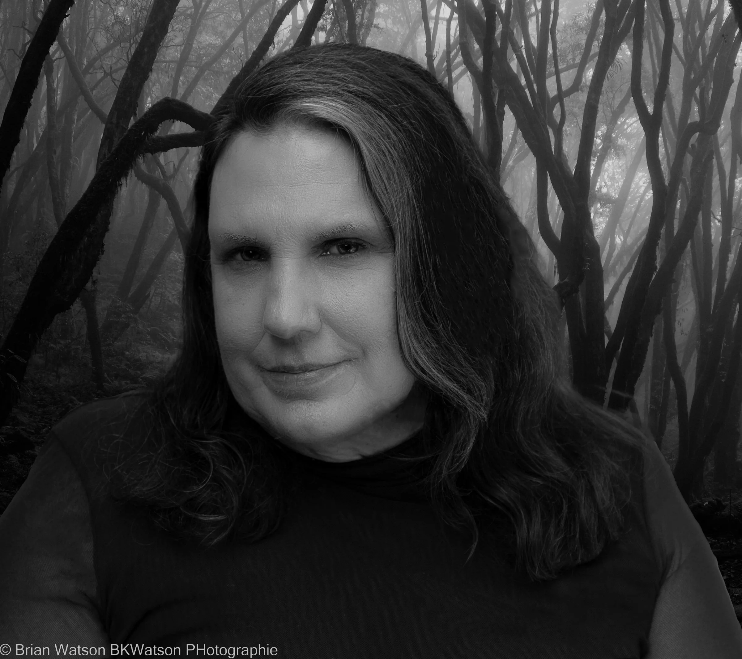 Black and white image of a woman smiling toward the camera in front of a background of bare tree limbs.
