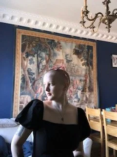 Image of a woman in black dress and shaved head looking out a window, framed by a tapestry hanging in the background.