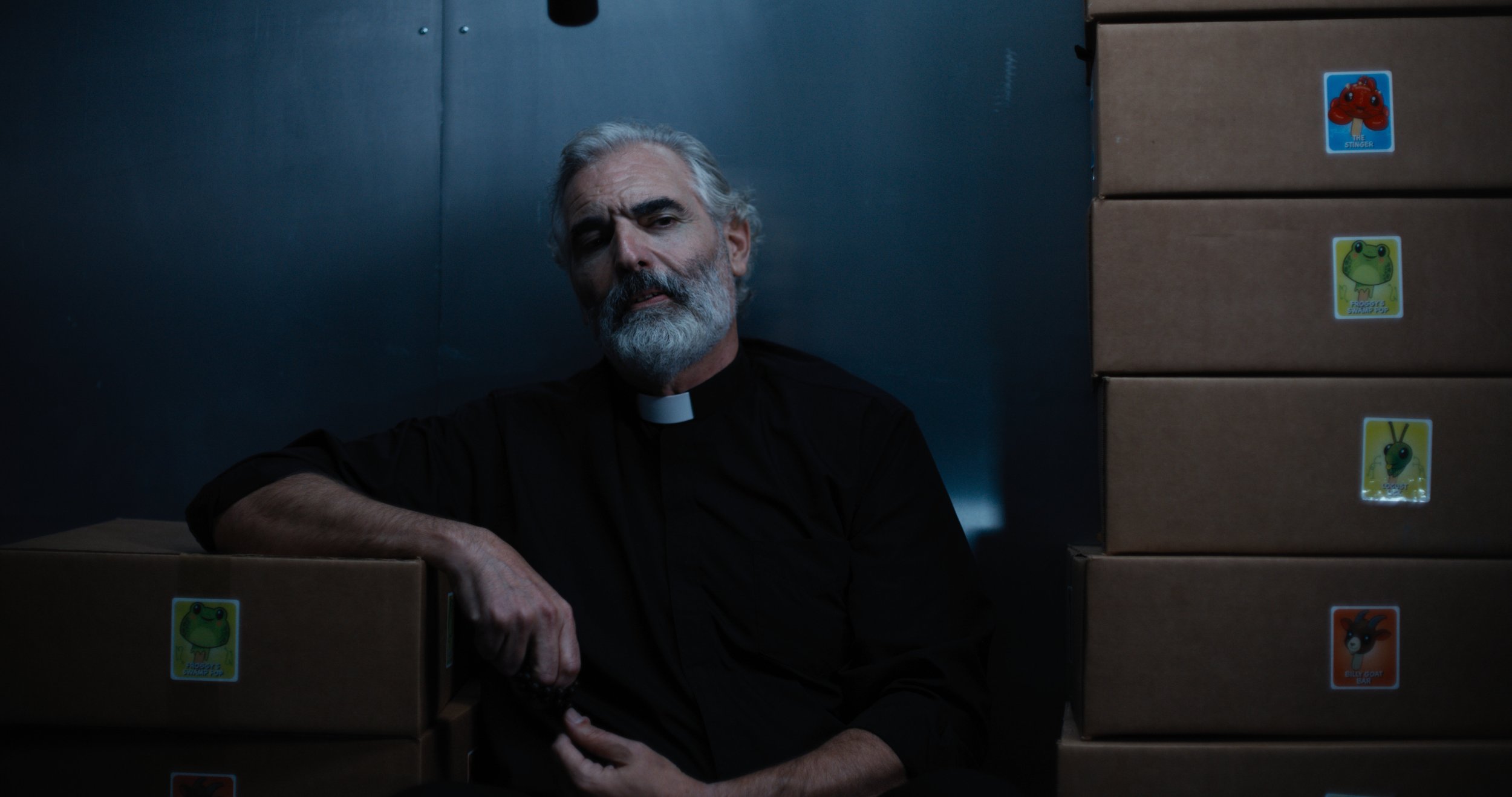 Older man with white hair and beard in a priest's garb, sitting in a dimly lit freezer surrounded by boxes of ice cream.