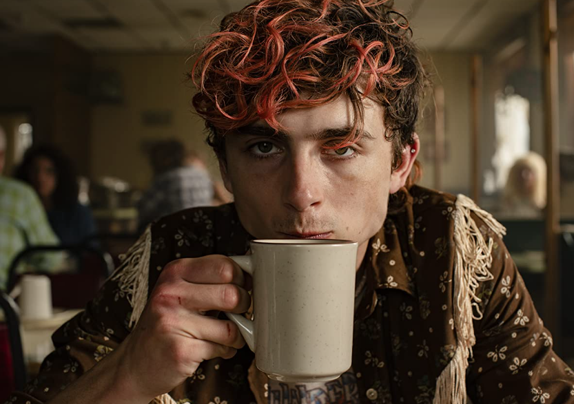 A young man with dyed orange hair sips on a hot cup of coffee. He looks directly at the camera, the hand holding the coffee cup is slightly bloodied.