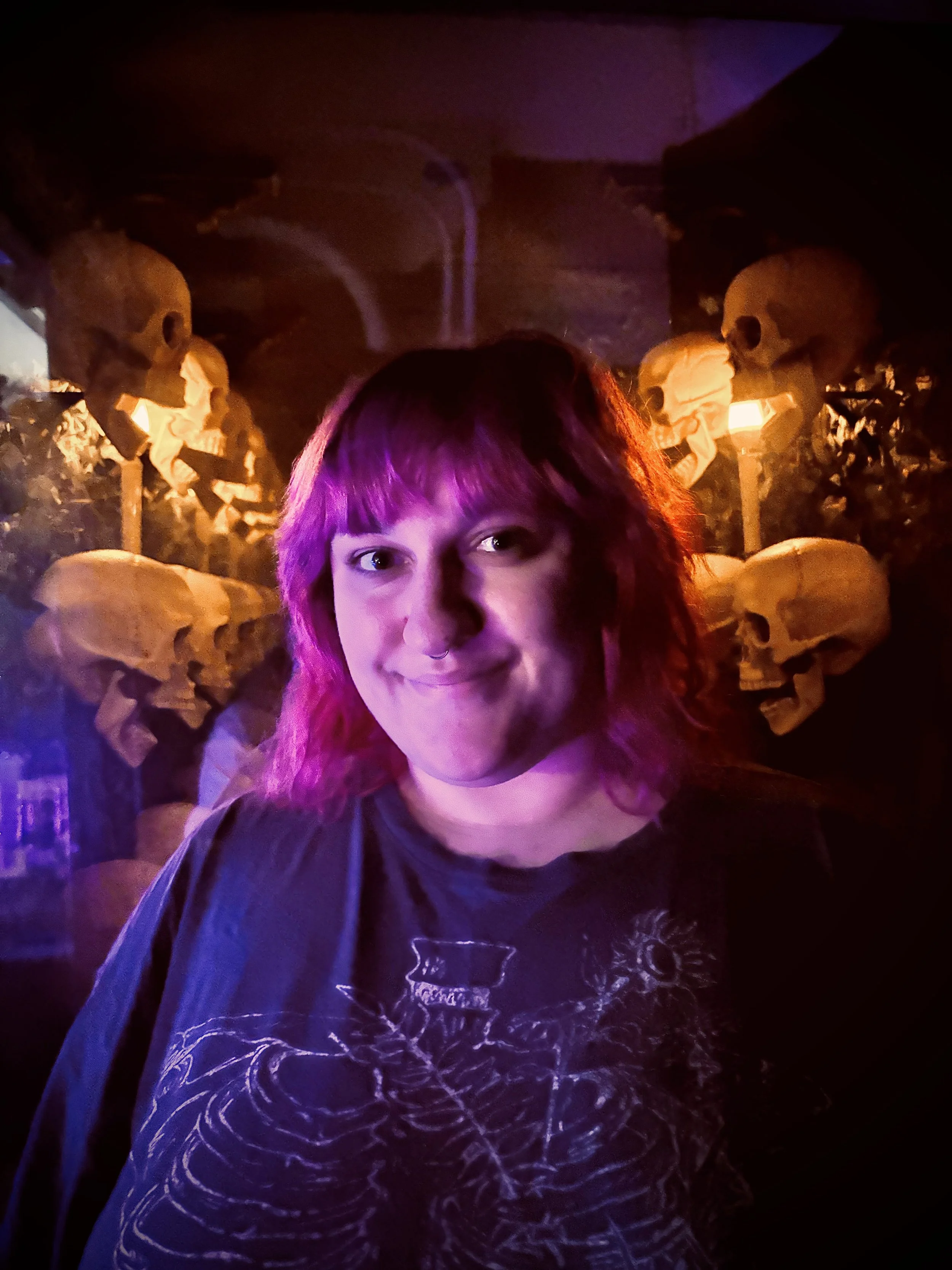 A pink-haired person smiles in a dimly lit room with skulls displayed behind them.