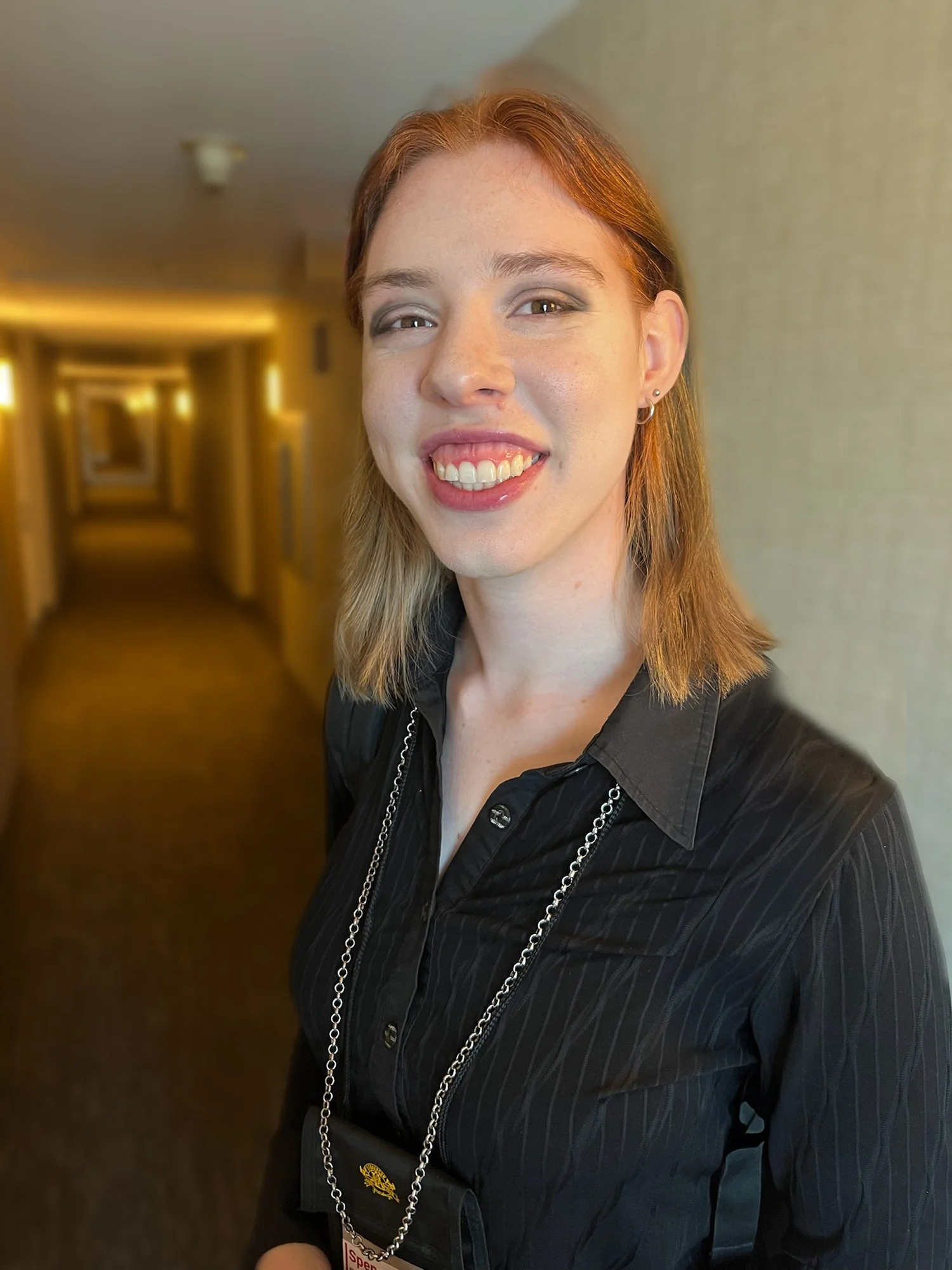 Image of a smiling person with a long hallway behind them. They wear a black collared shirt with a silver chain around their neck.