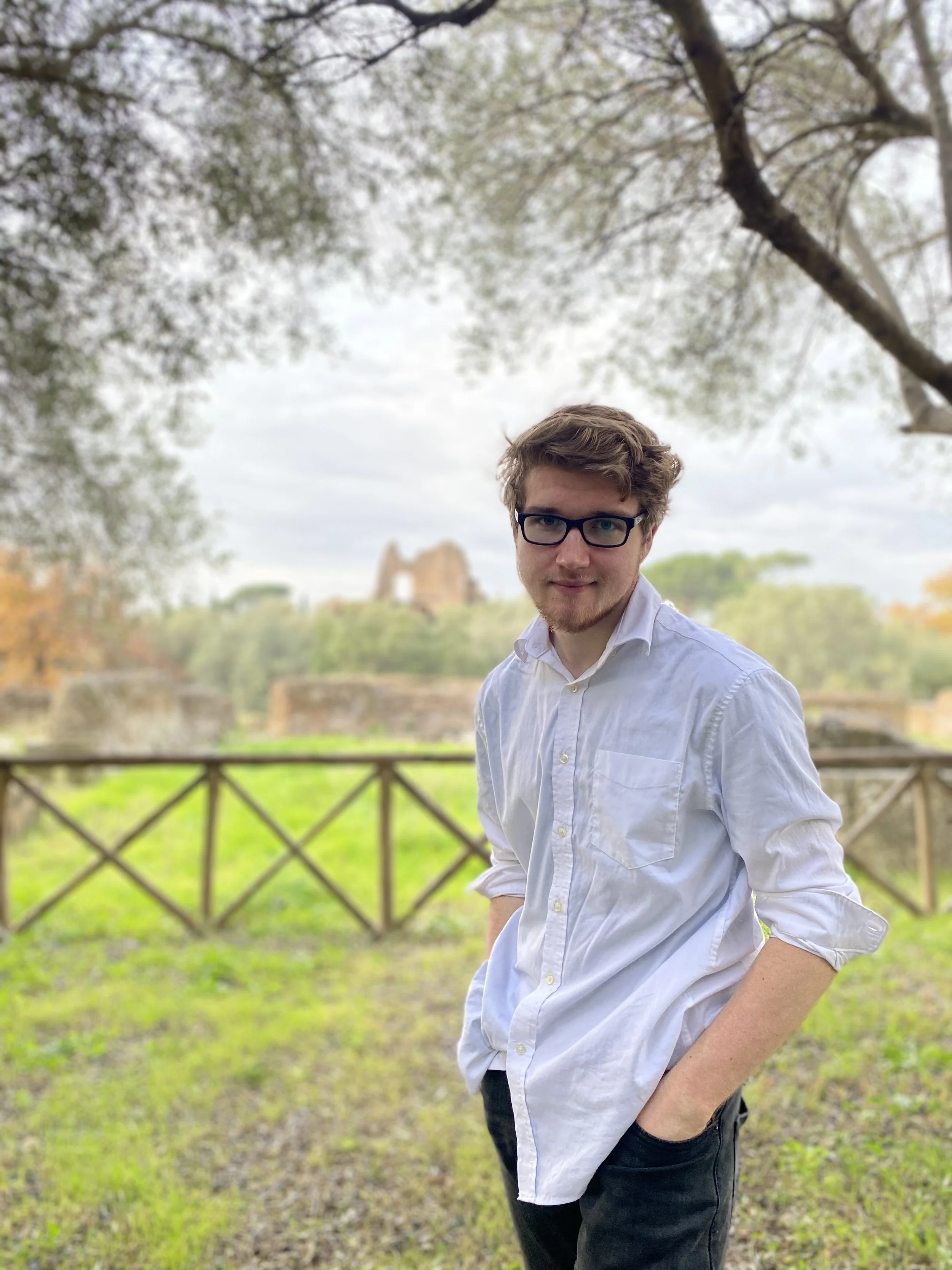 Young man wearing a white, untucked button-down shirt with hands in pocket. Standing in a green field.