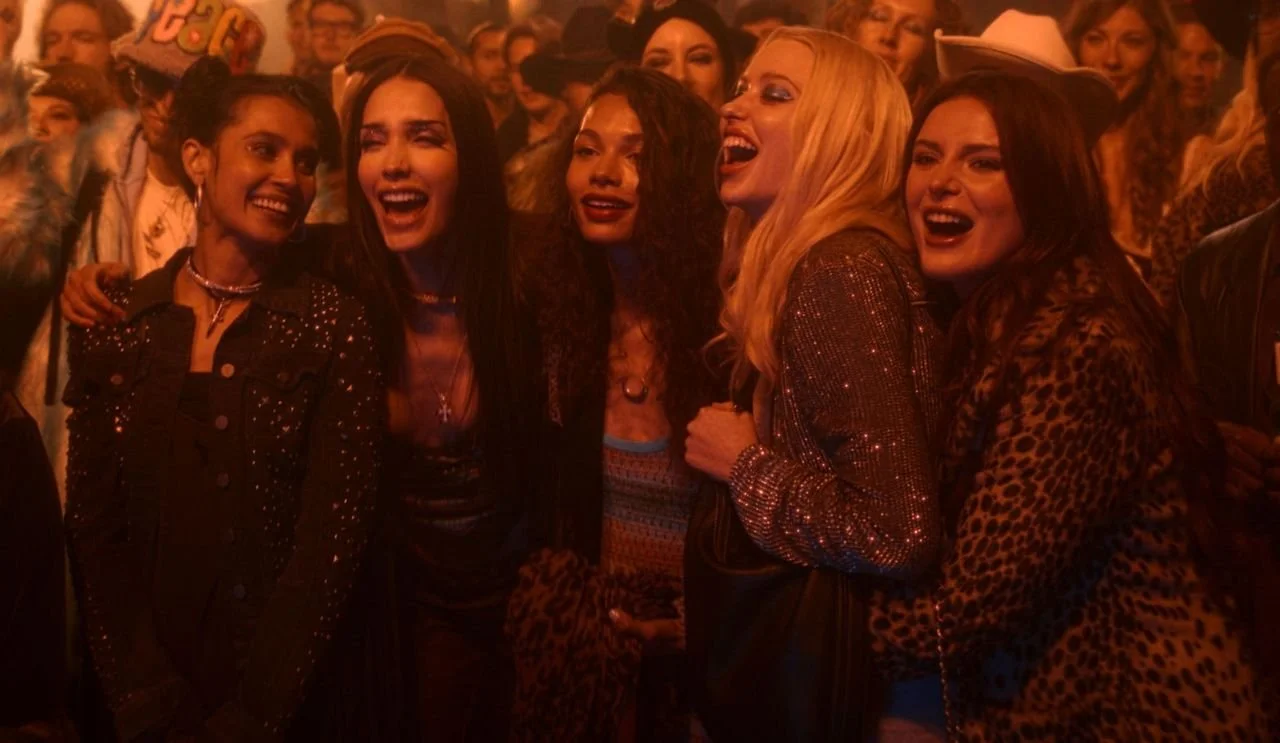Five women stand arm-in-arm in a red-tinged image with partyers in the background.