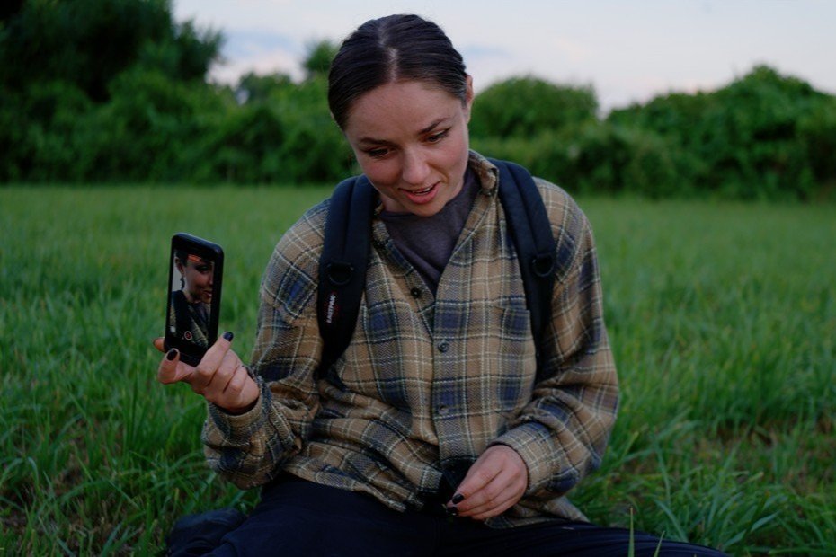 A girl wearing a flannel shirt and backpack sits in a field of green grass with her phone in her hand, recording a video of herself.