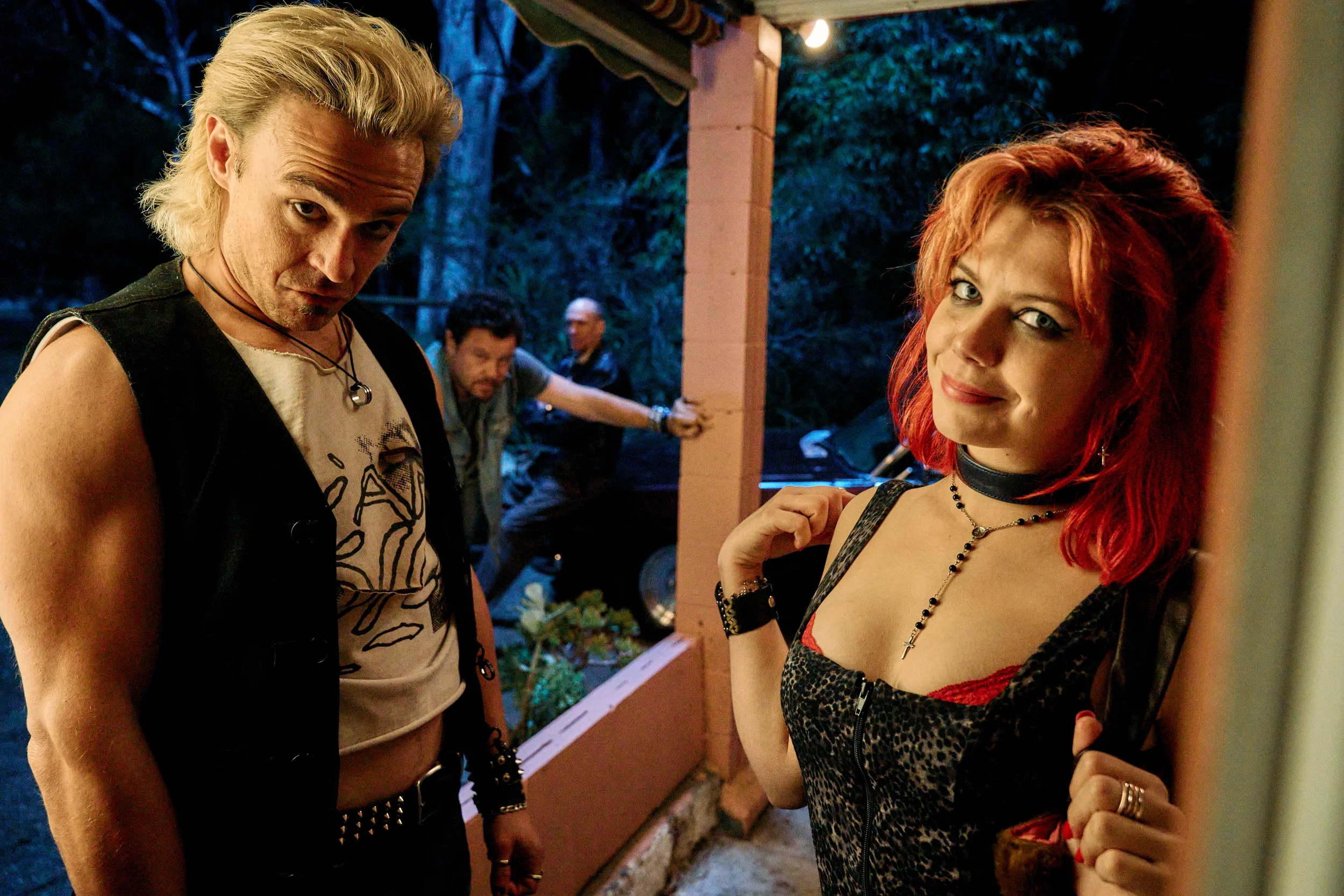A man with bleached, slicked back hair wearing a cutoff tee and vest stands to the left, a dyed-red haired woman in leopard print stands on the right. They peer in through a doorway with menacing grins on their faces.