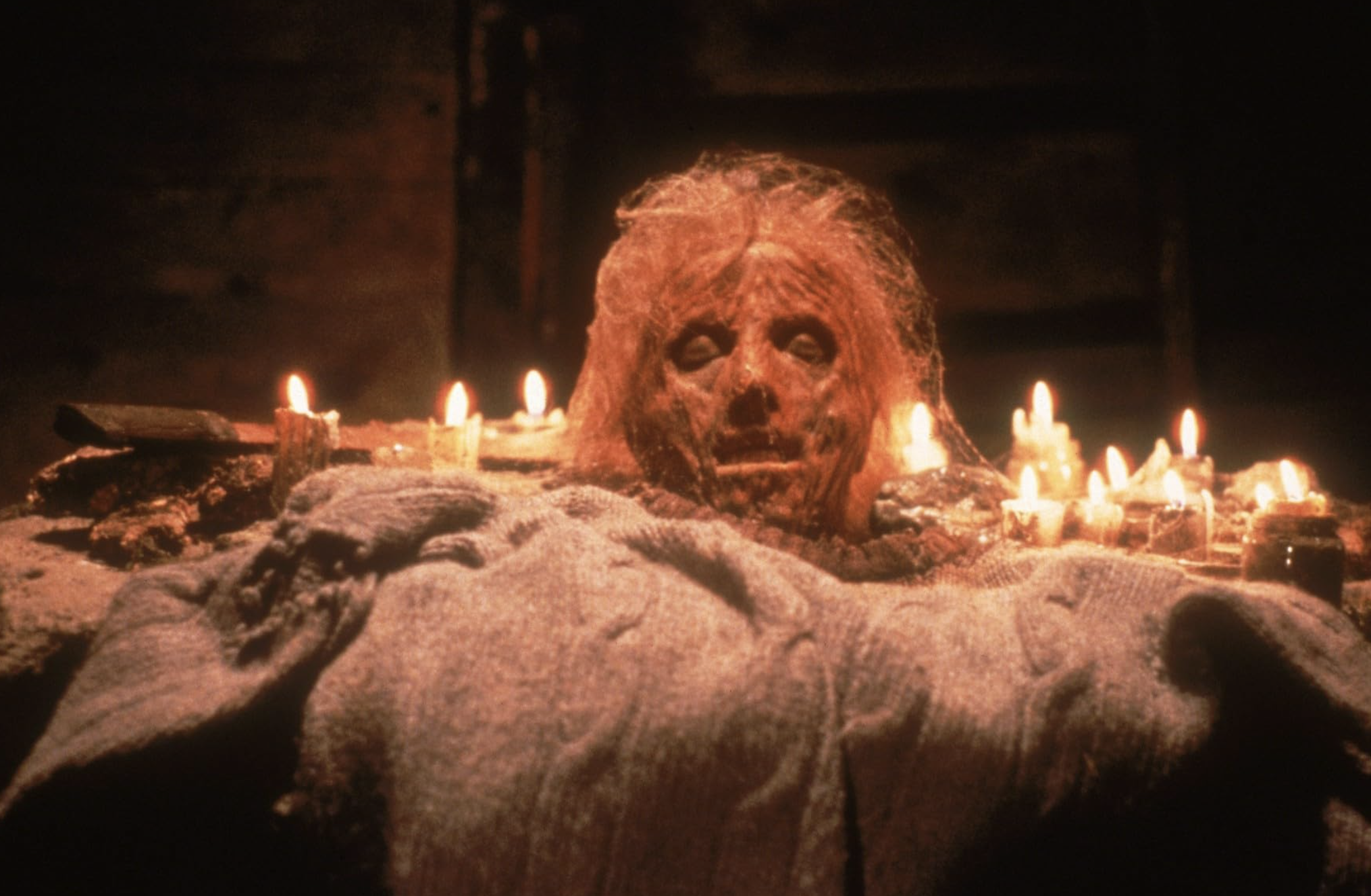 A severed and rotting head sits atop an altar cloth surrounded by lit candles.