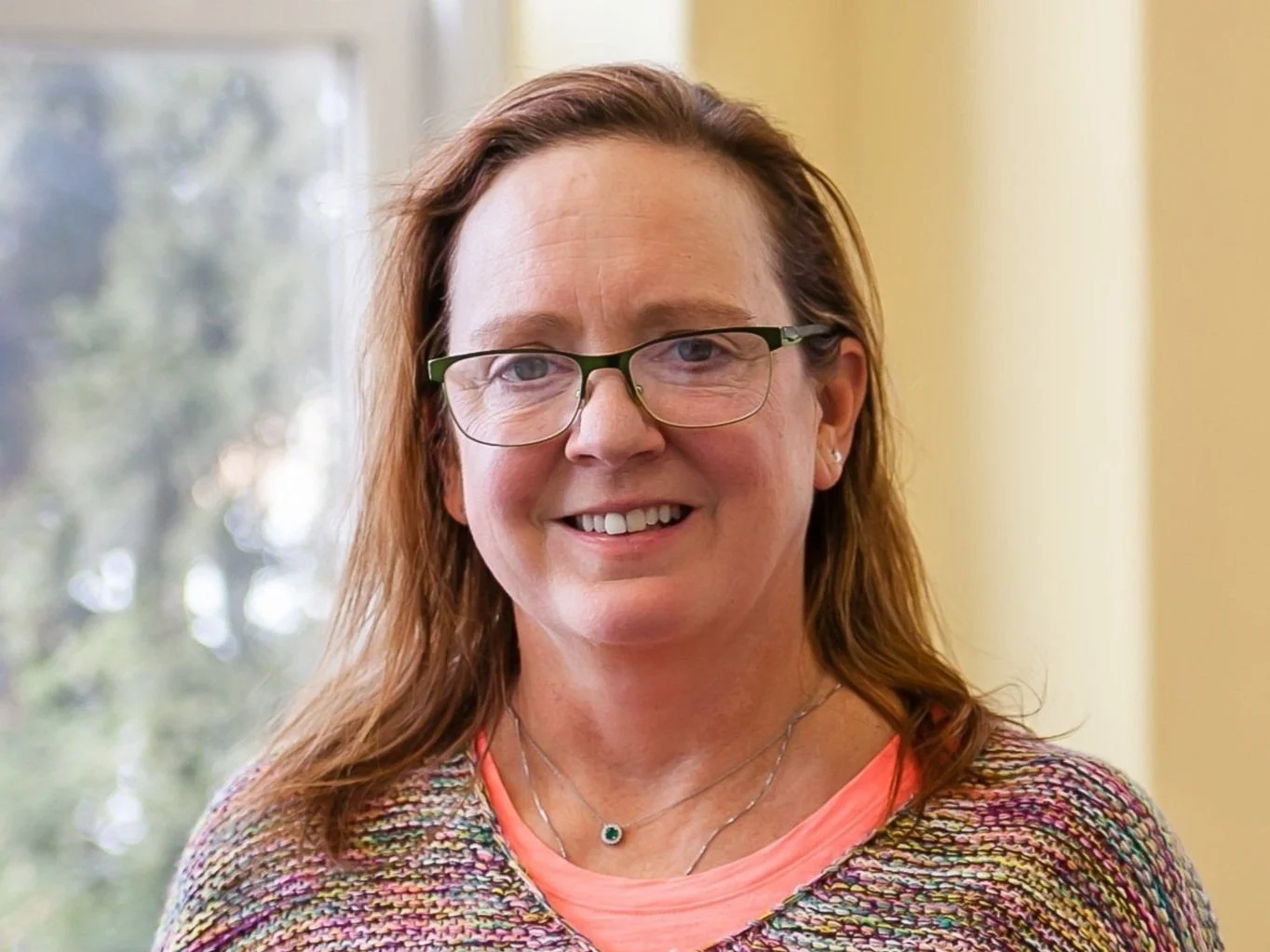 A woman with reddish brown hair wearing glasses, earrings, a multicolored knit top, and a pink shirt underneath. She is smiling and standing indoors near a window.