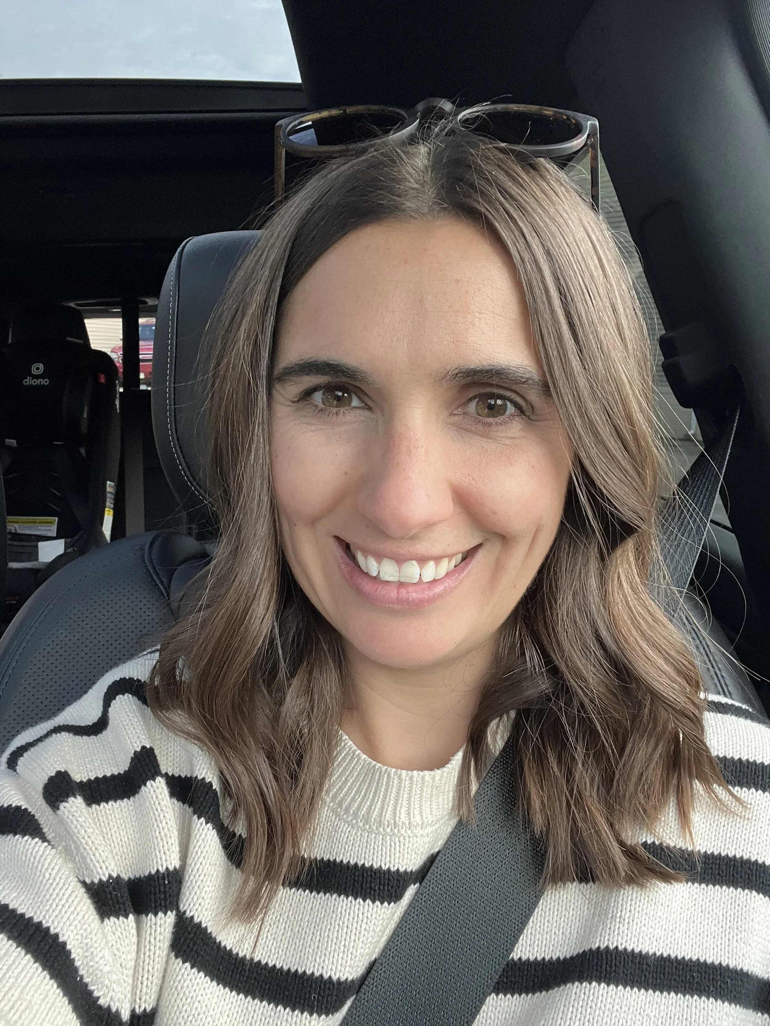 A woman with long, wavy brown hair smiling inside a car, wearing a black and white striped sweater and sunglasses on her head, with a baby car seat in the backseat.