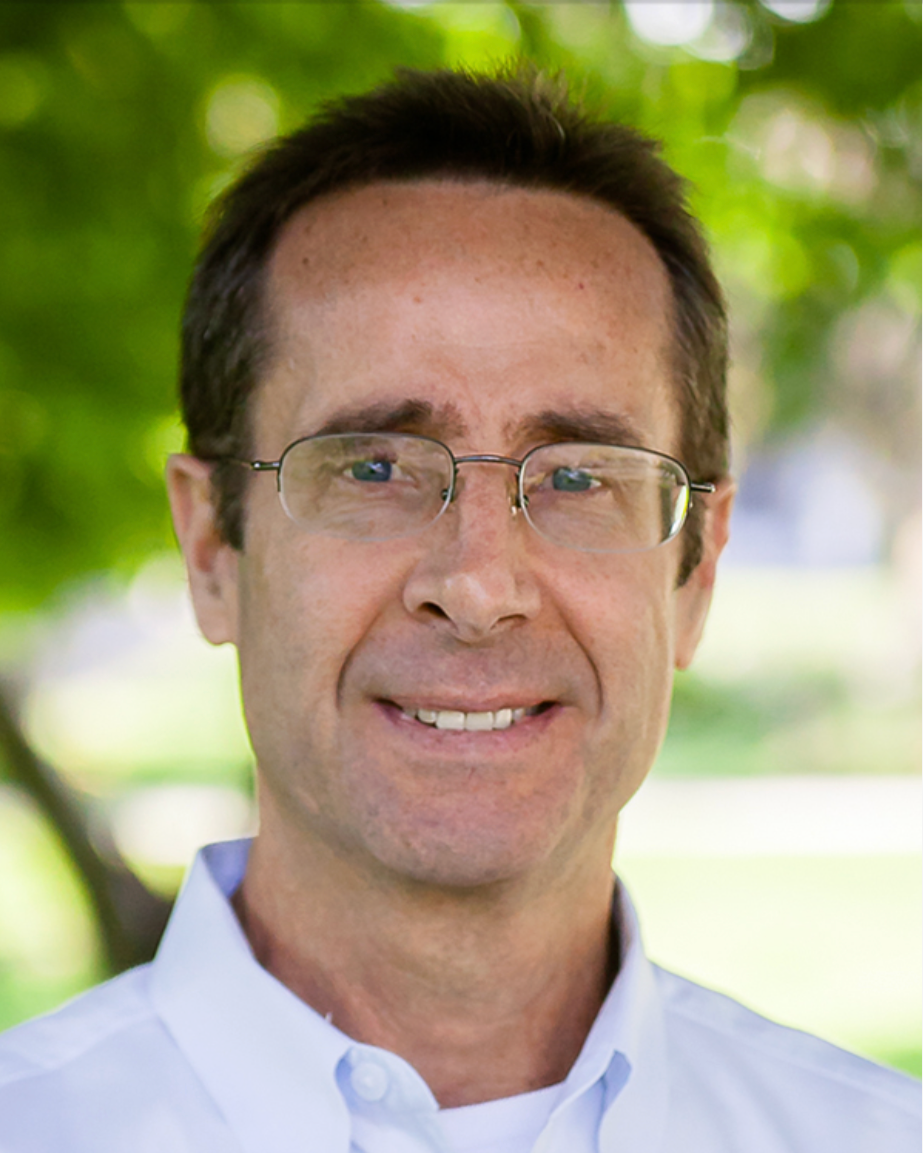 A man wearing glasses and a white collared shirt outdoors, smiling with trees and greenery in the background.