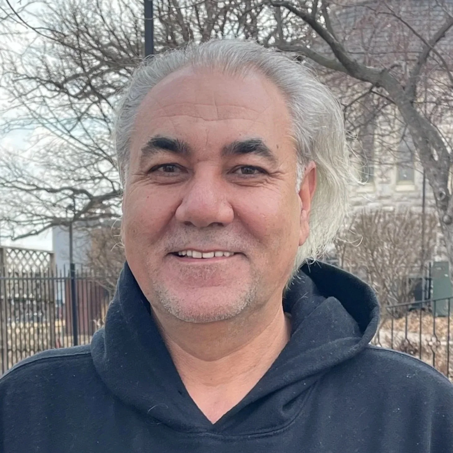 Close-up of a smiling middle-aged man with gray hair, wearing a dark hoodie, outdoors with leafless trees and a metal fence in the background.