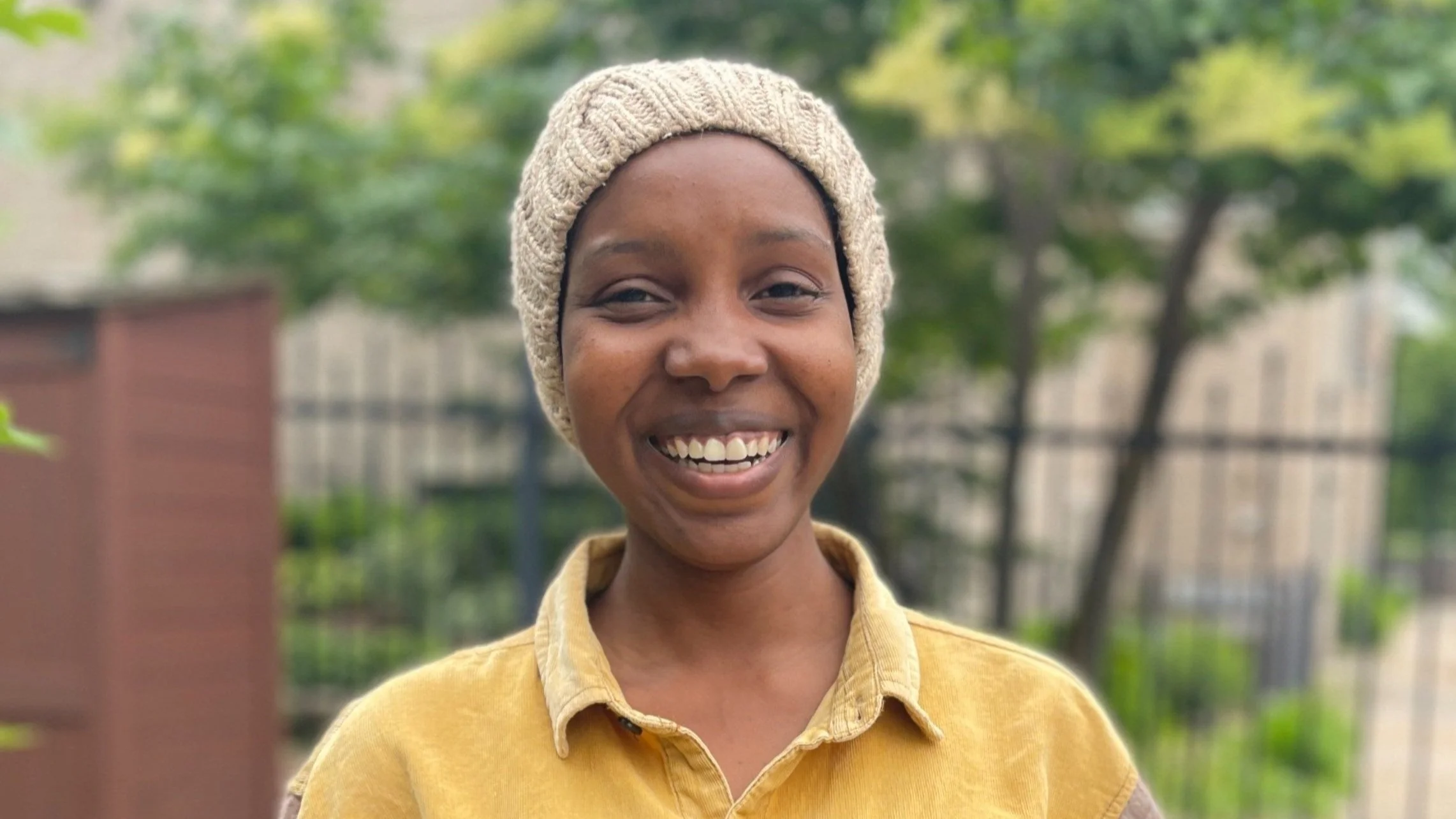 A smiling woman wearing a knitted beige hat and a yellow shirt outdoors with trees and a fence in the background.