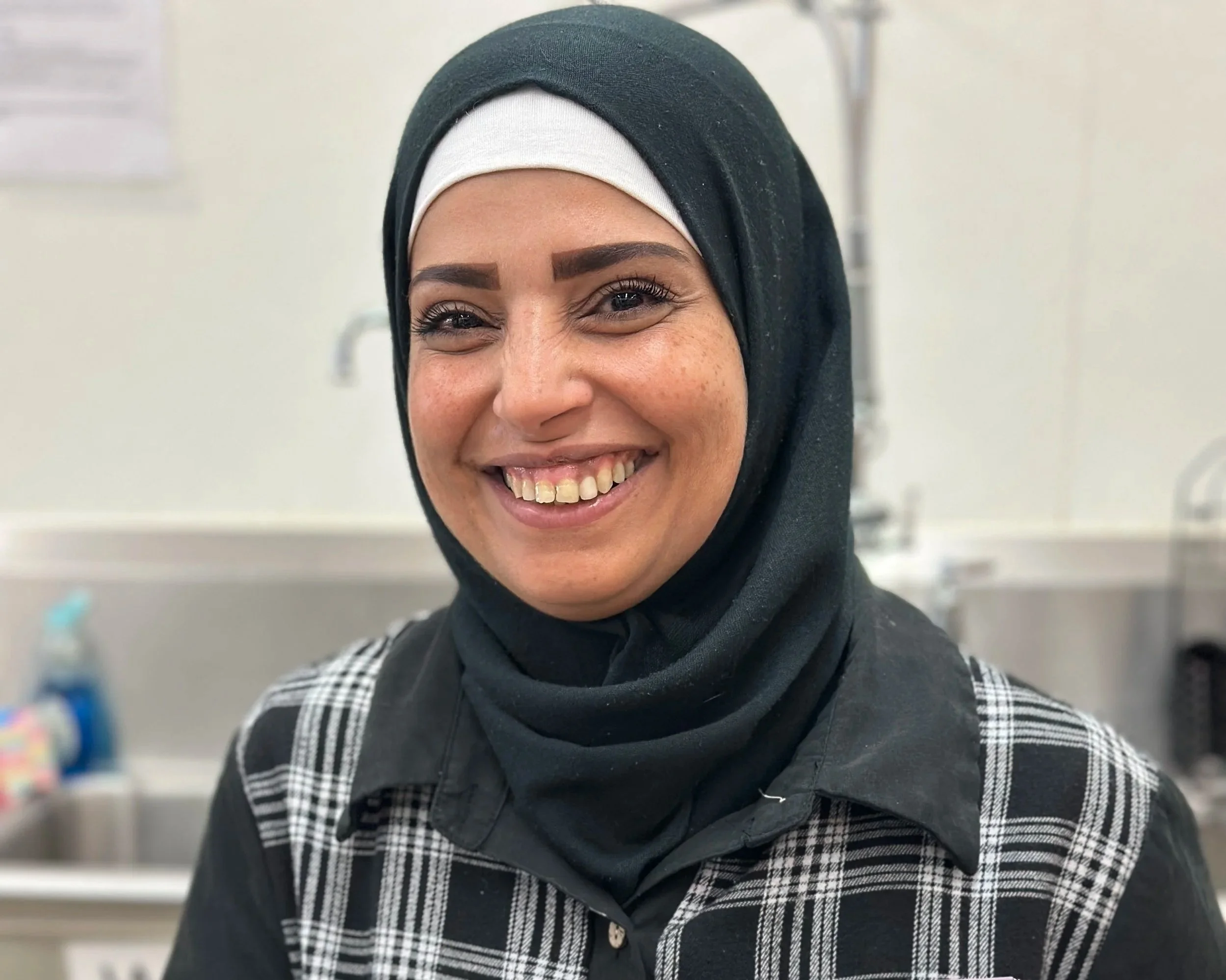Close-up of a smiling woman wearing a black hijab with a white inner lining, a gray and black patterned shirt, and makeup. The background appears to be a bright indoor setting.