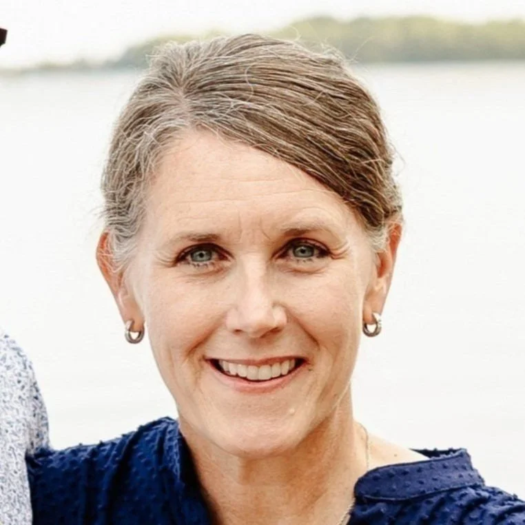 A smiling woman with short brown hair, wearing earrings and a navy blue shirt, outdoors near a body of water.