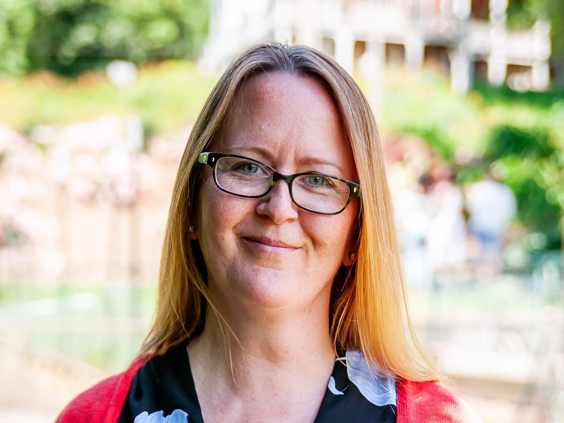 Close-up of a smiling woman with glasses and long, light brown hair standing outdoors with blurred green trees and buildings in the background.