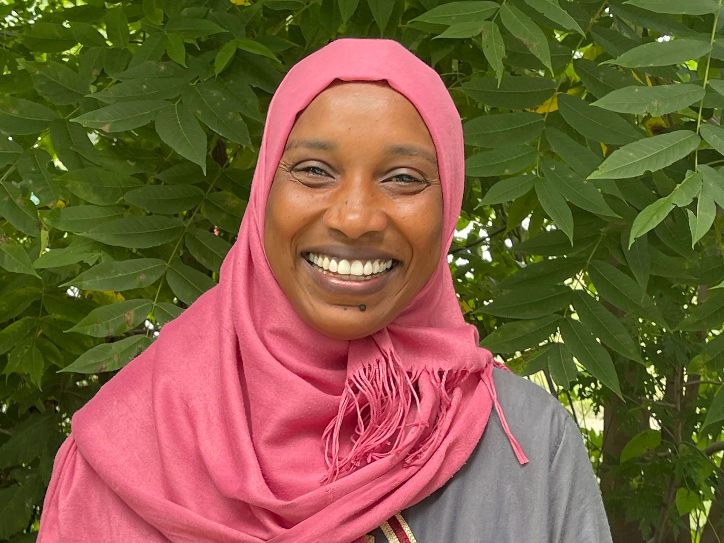 A woman smiling with a pink headscarf, sitting outdoors near lush green leaves.