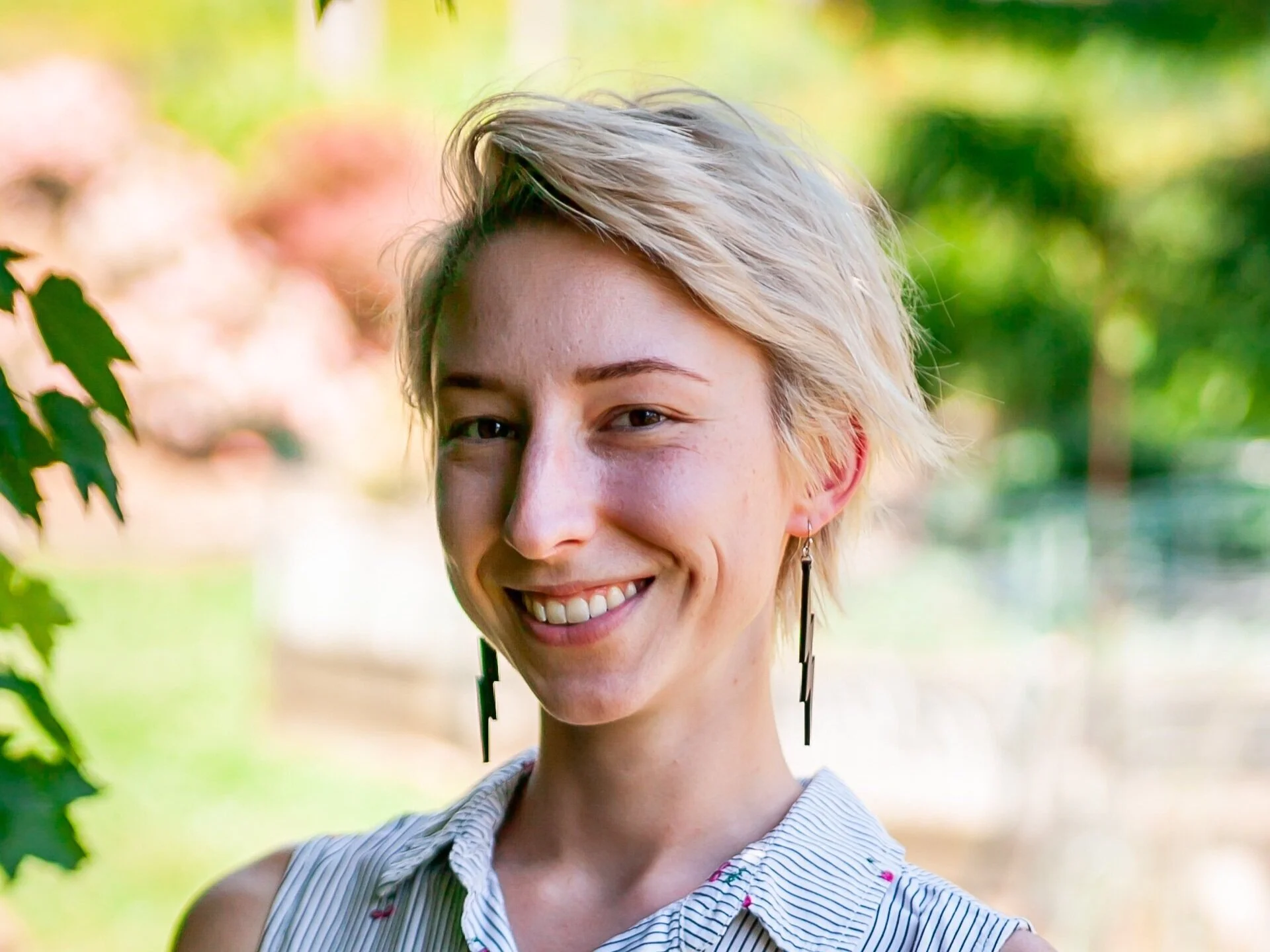 Close-up of a smiling woman with short blonde hair, wearing earrings and a striped sleeveless shirt, outdoors with green and pink blurred background.