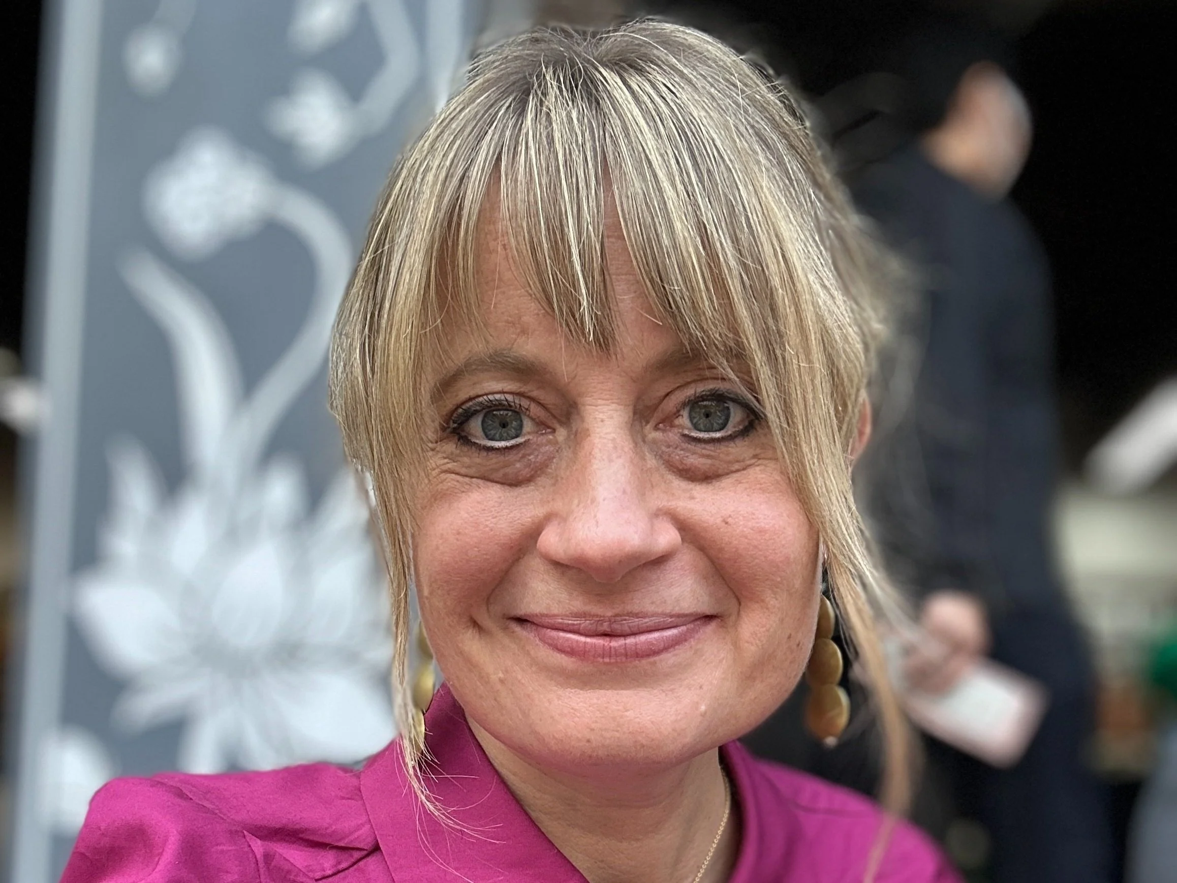 A close-up of a woman with short blonde hair, gray eyes, and a pink top, smiling at the camera.