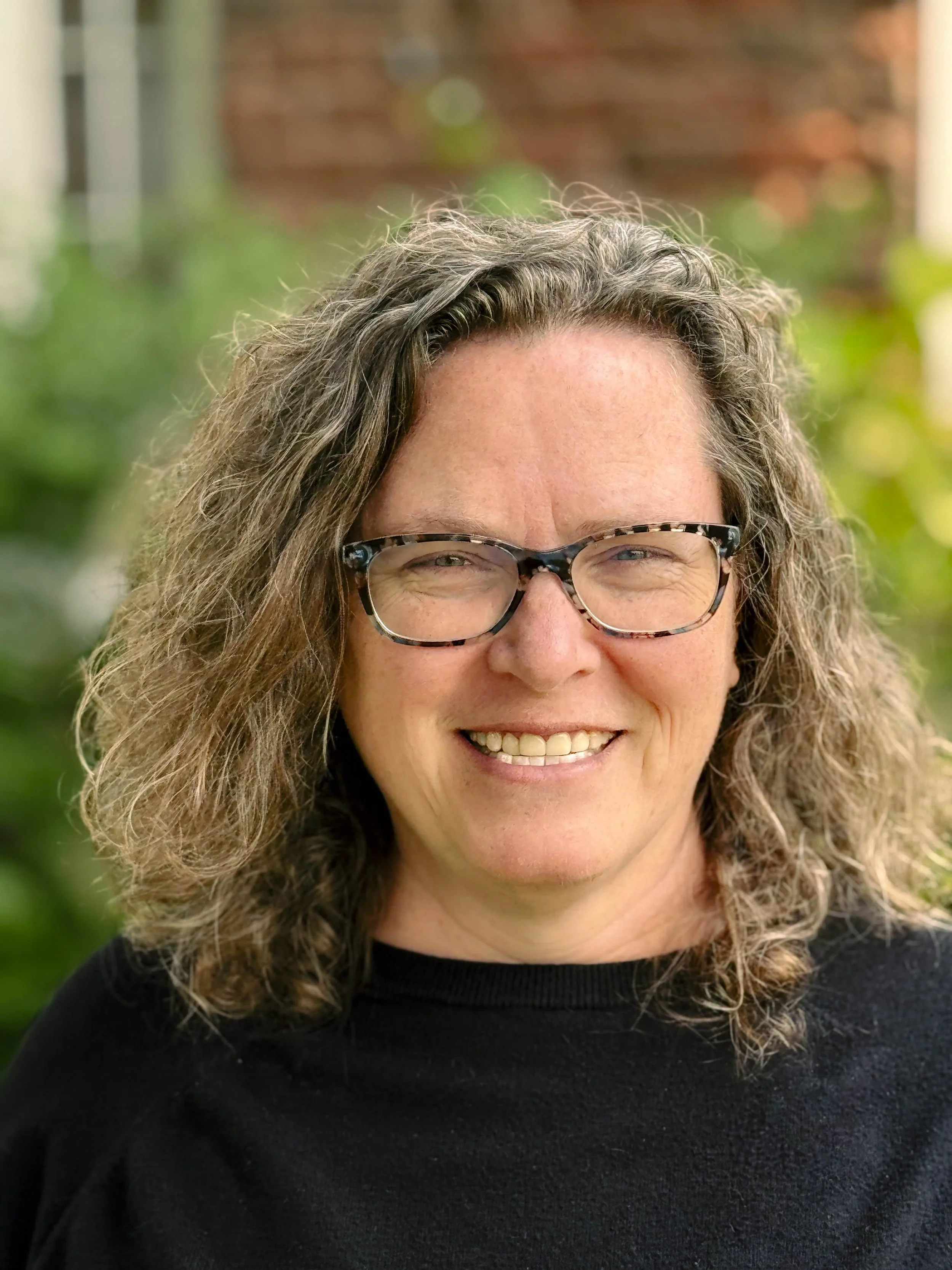 Close-up of a smiling woman with wavy, shoulder-length gray hair and styled glasses, wearing a black top, outdoors with blurred greenery in the background.