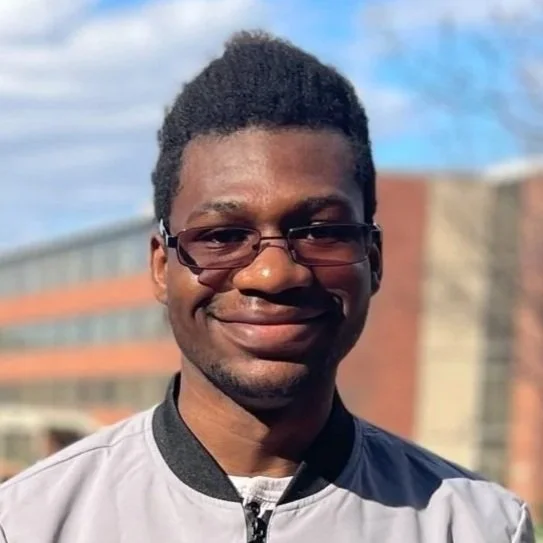 A young man with glasses and short curly hair, smiling outdoors on a sunny day with a building and blue sky in the background.