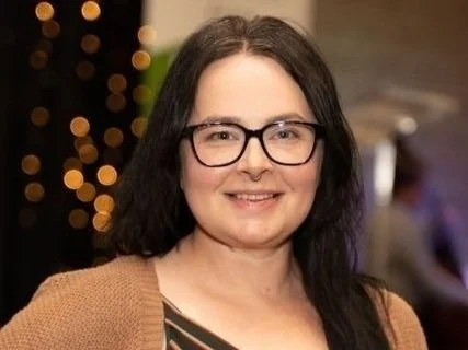 A woman with shoulder-length dark hair and glasses smiling in an indoor setting with blurred lights in the background.