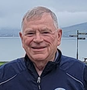 A smiling elderly man with short gray hair wearing a navy blue jacket, standing outdoors near a body of water with a cloudy sky in the background.