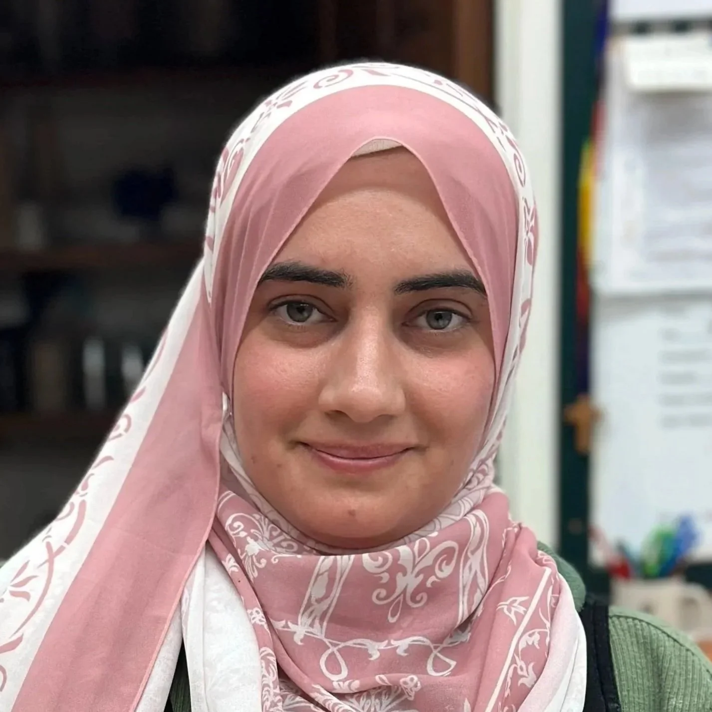 A woman with light skin and gray eyes wearing a pink and white patterned hijab, smiling.