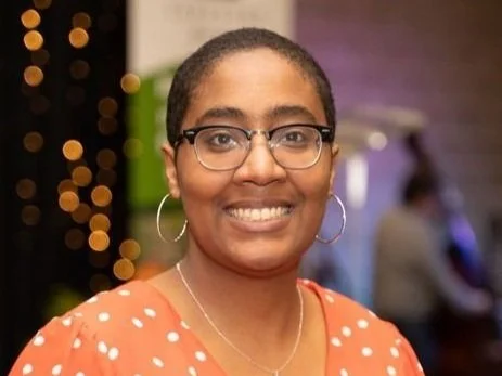 A woman with glasses and hoop earrings smiling at a social event with decorative lights in the background.