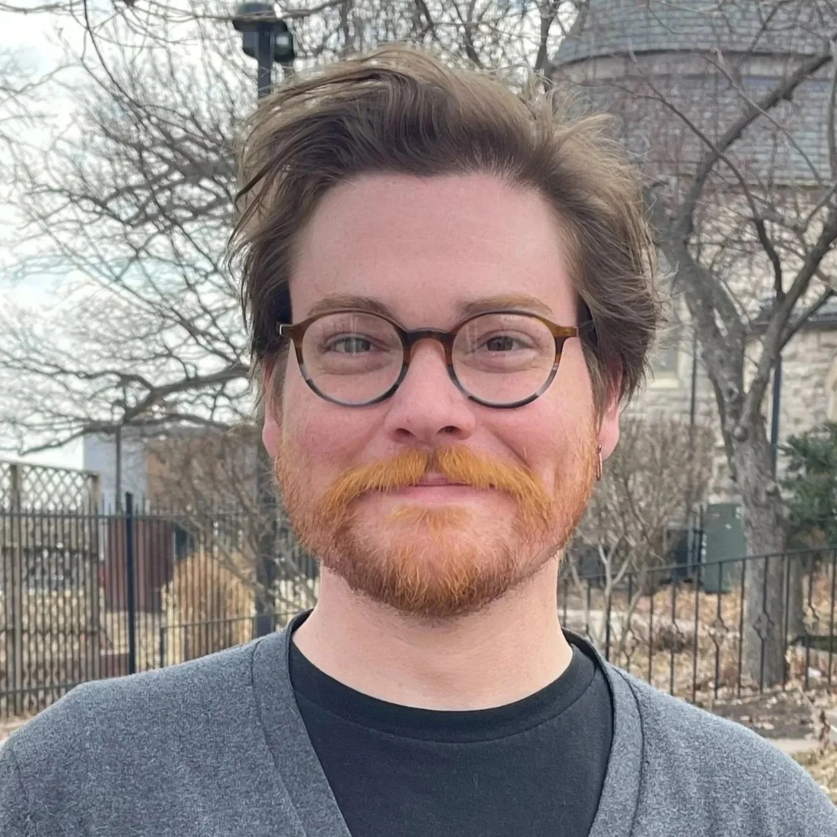 Close-up of a smiling man with glasses, reddish beard and hair, standing outdoors with leafless trees and buildings in the background.
