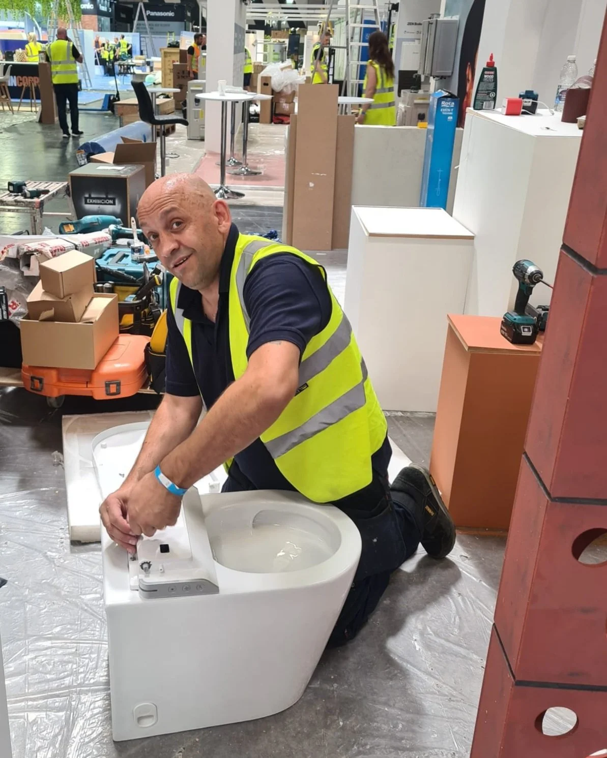 A man wearing a yellow safety vest kneeling on the floor installing a white bidet. He is surrounded by tools and boxes, with other workers and display booths in the background at what appears to be a trade show or showroom.