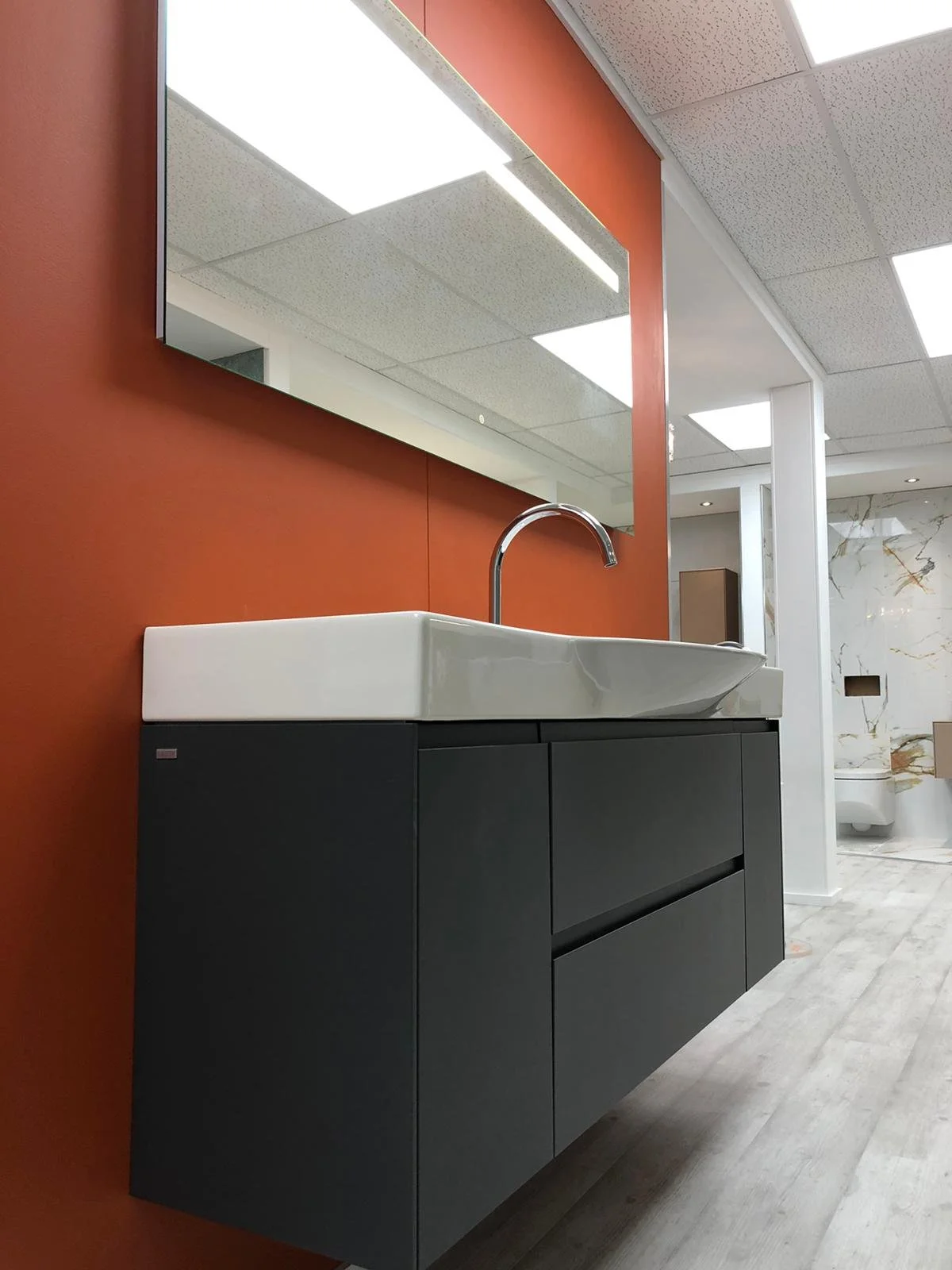 Modern bathroom vanity with a black cabinet, white sink, and chrome faucet, set against a rust-colored wall with a large mirror above, in a contemporary bathroom with marble accents.