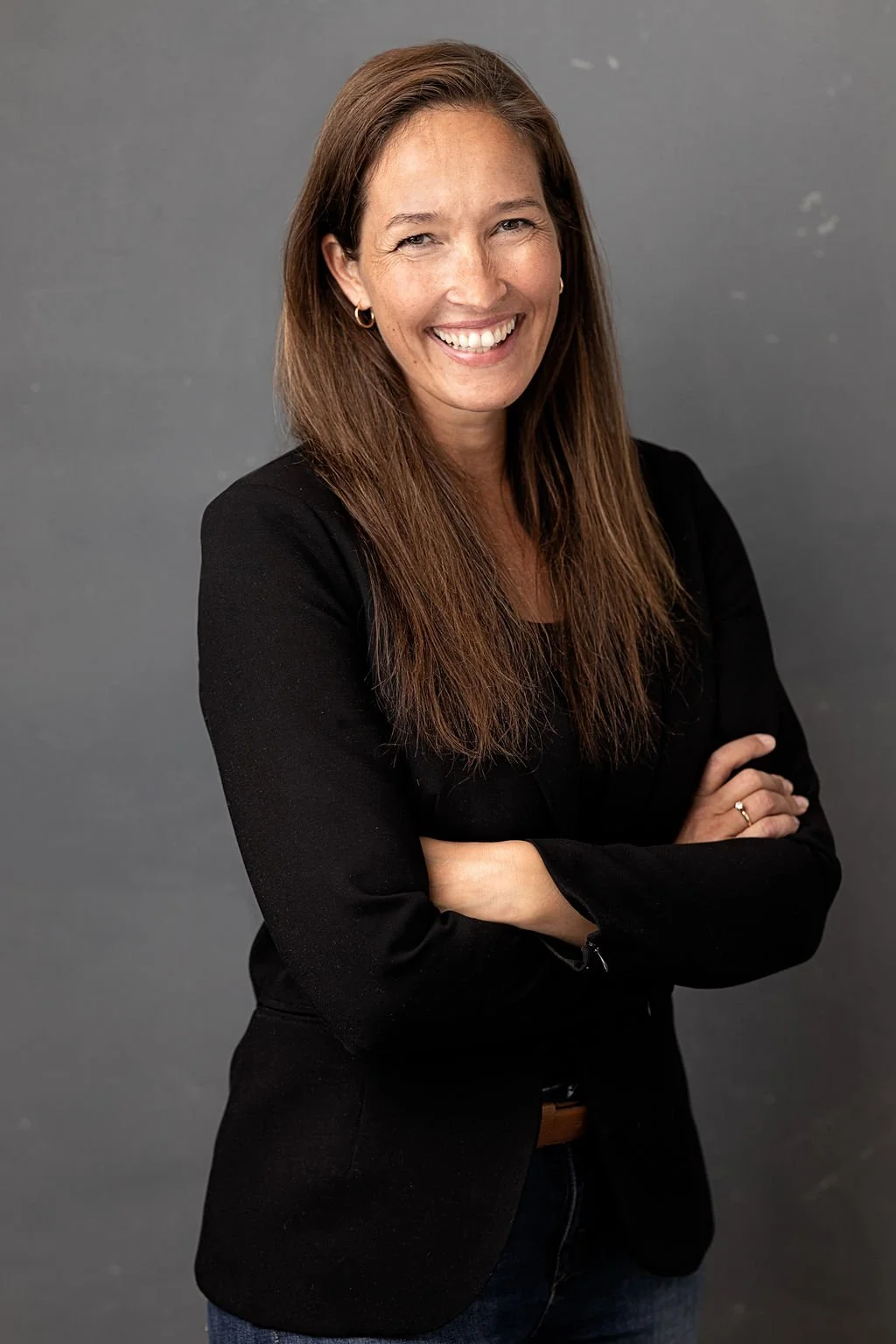 A woman, Camilla Brox, with long brown hair smiling and standing with arms crossed, wearing a black blazer and dark jeans against a gray background.