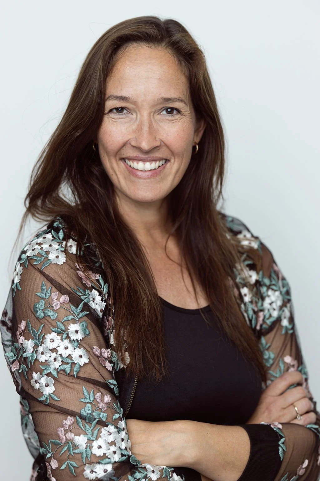 A woman with long brown hair smiling at the camera, wearing a black top and a floral embroidered sheer jacket, standing against a plain light background.
