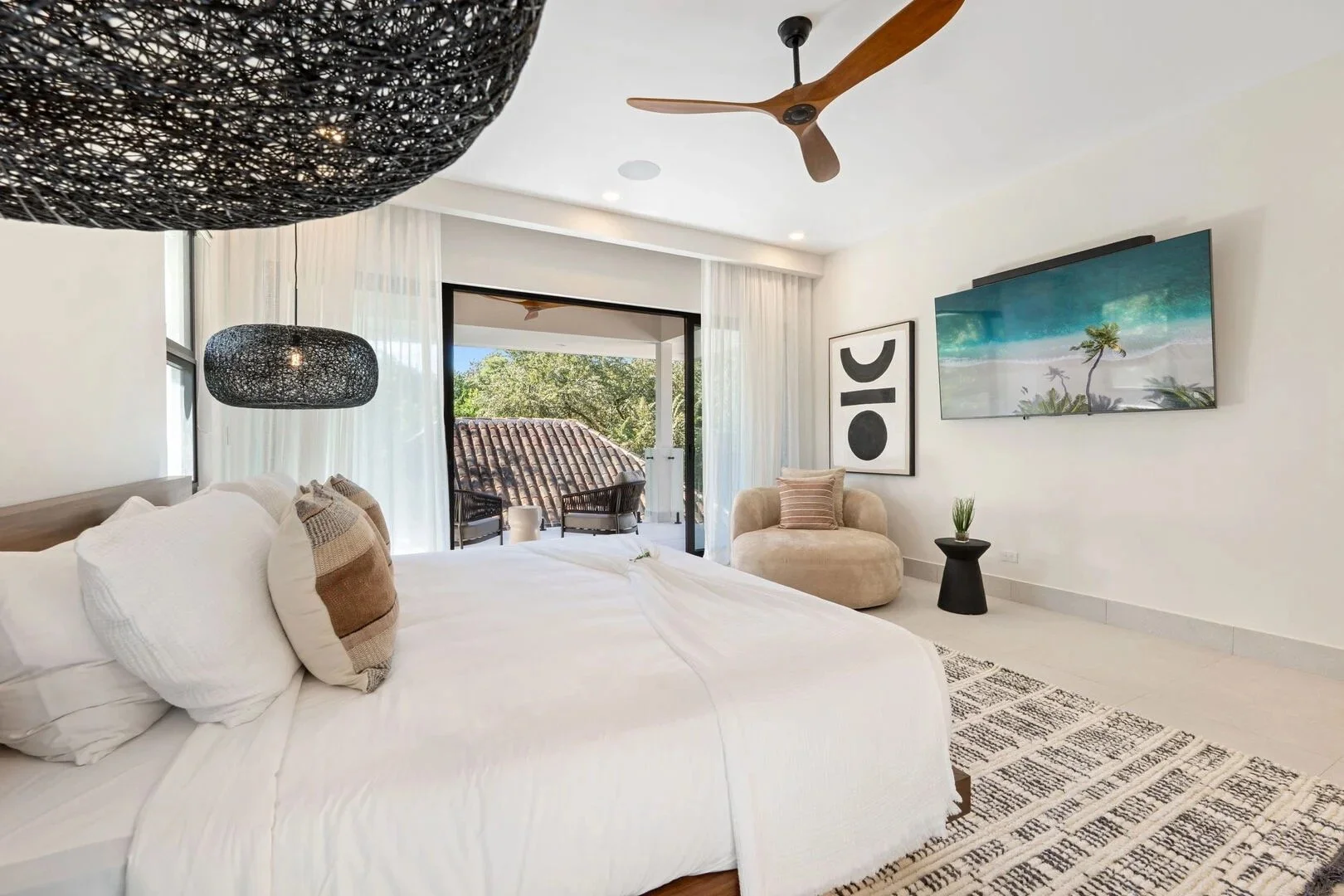 Modern bedroom with white bedding, balcony doors, outdoor seating, black woven pendant lights, wall art, and a wall-mounted TV.