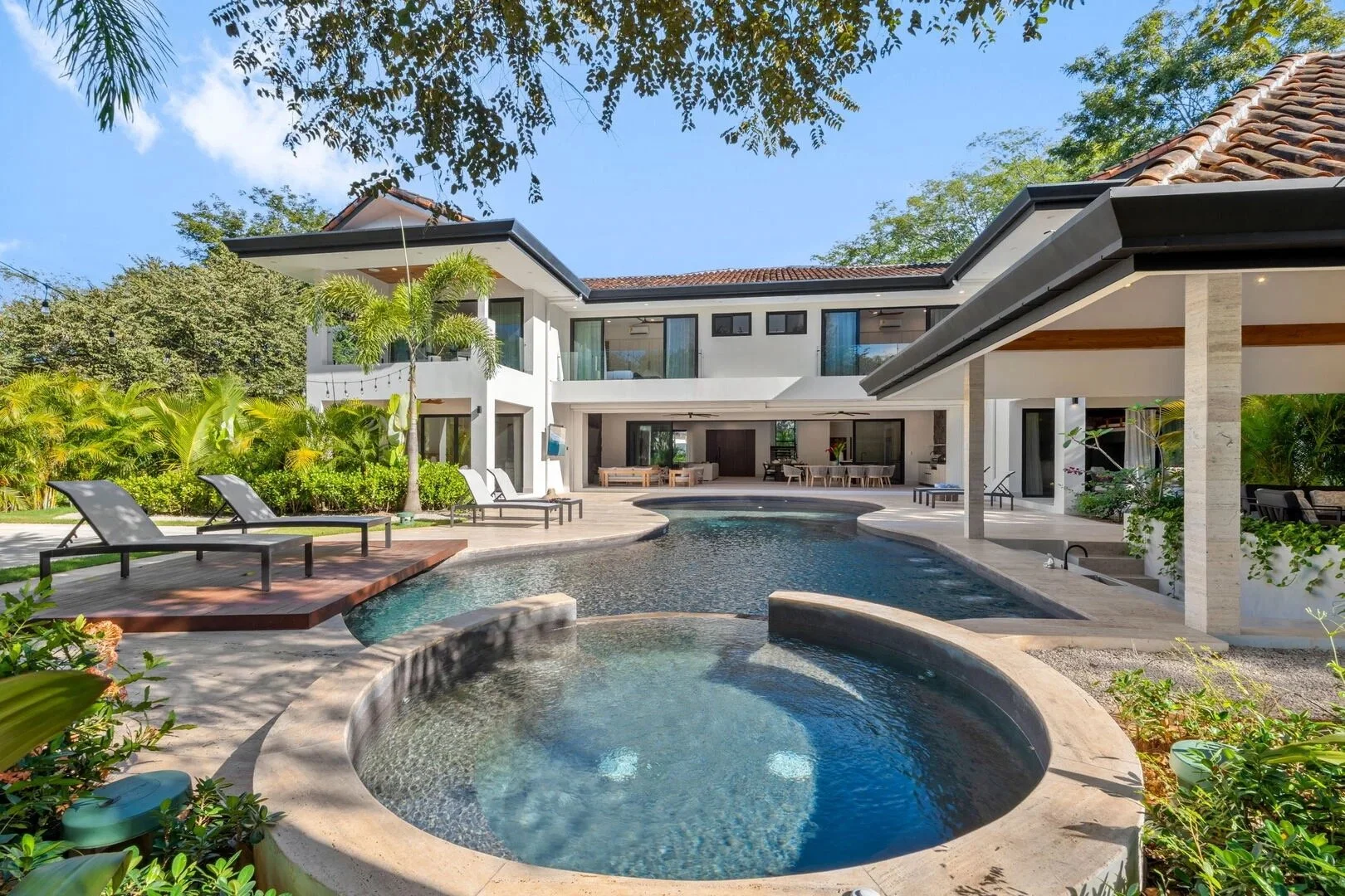Luxury modern house with outdoor pool, poolside lounge chairs, lush landscaping, and a covered patio on a sunny day.