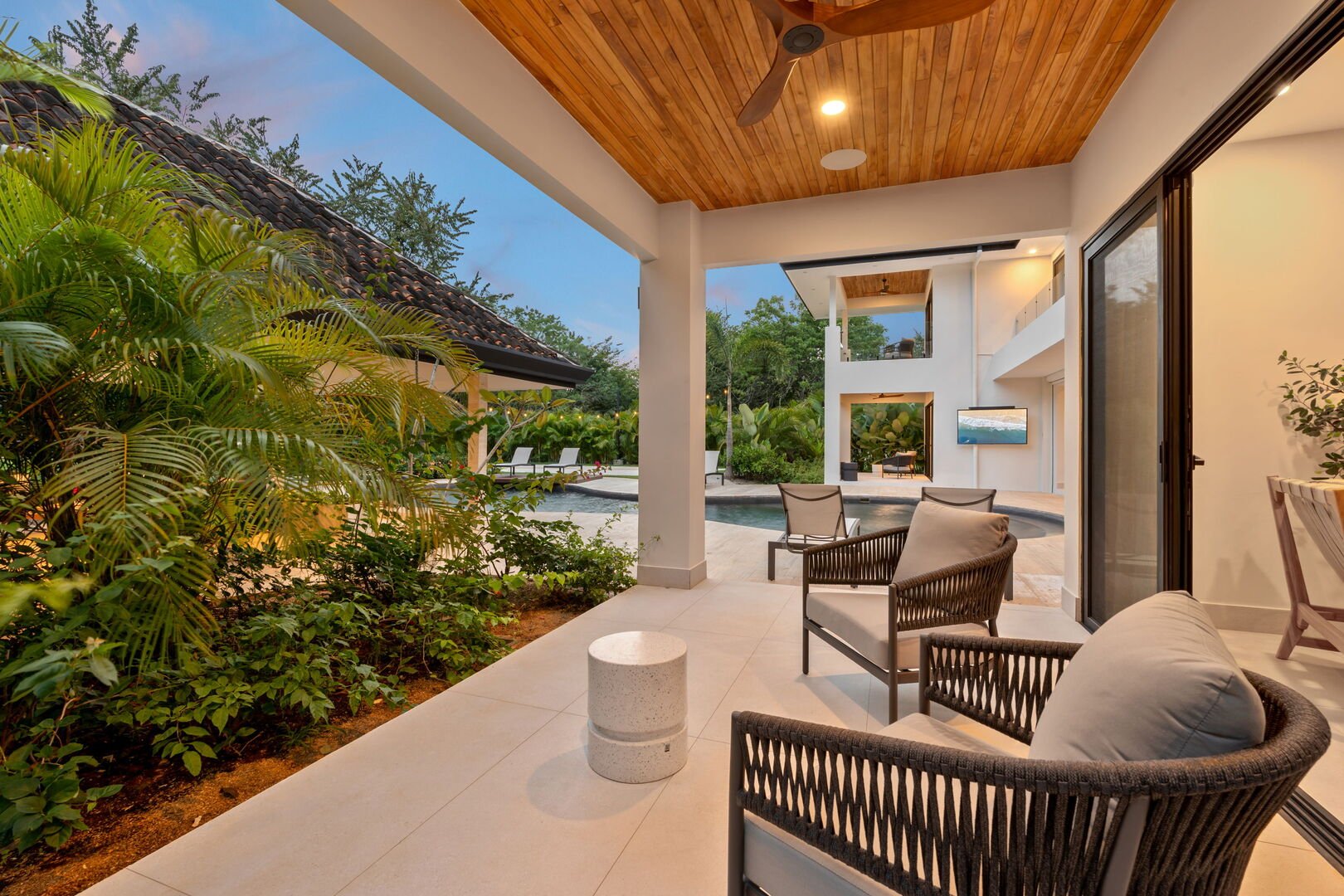 Climate-controlled outdoor patio with modern furniture, view of a pool, lush greenery, and a two-story house with large windows, during evening.