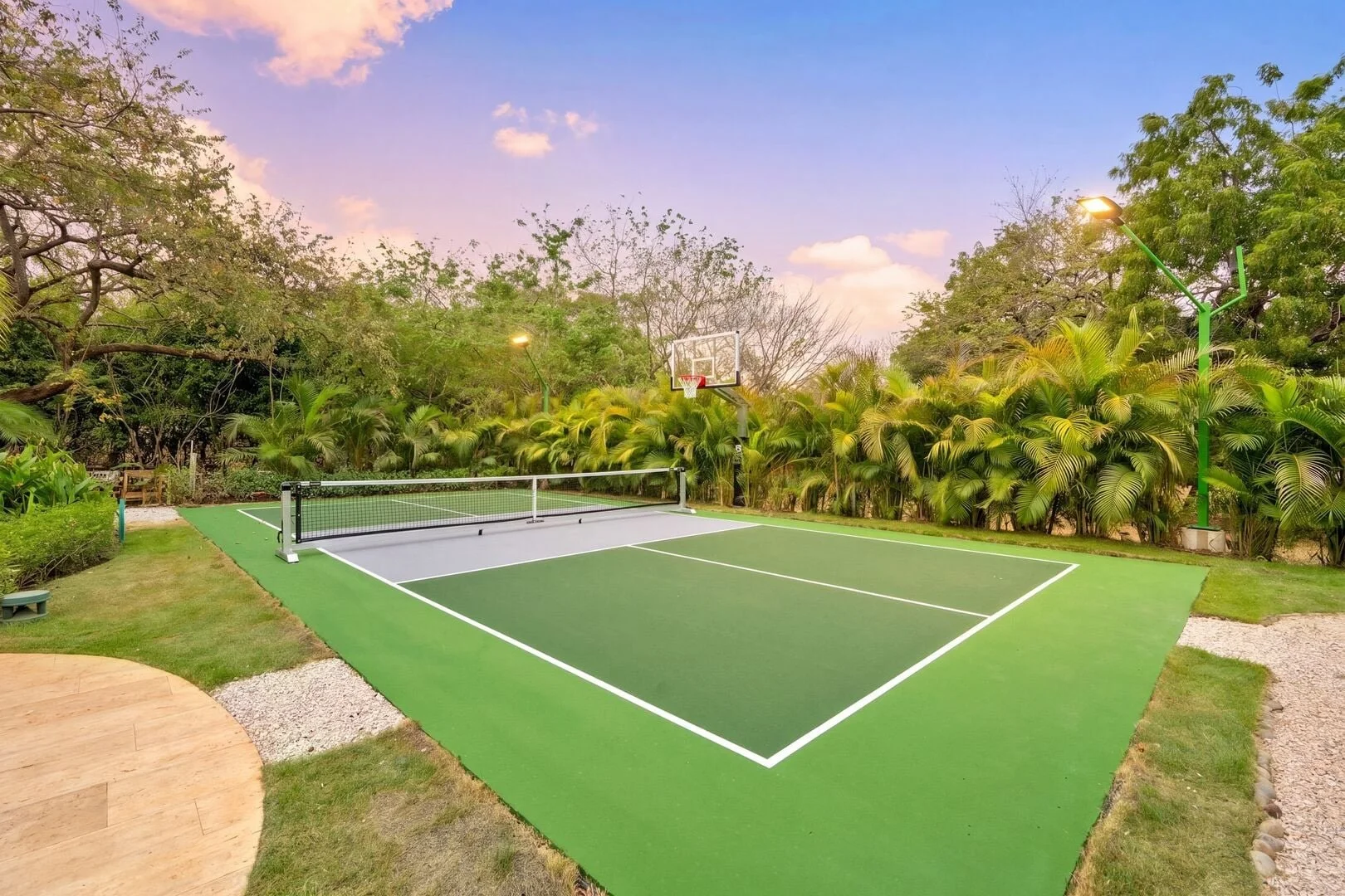 An outdoor sports court featuring a green tennis or pickleball court with white boundary lines, a net, a basketball hoop, and surrounded by lush greenery and trees, with a sky displaying a pinkish hue.