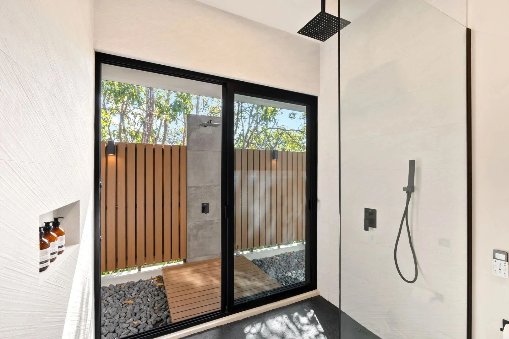 Modern bathroom with a walk-in shower, black fixtures, and a large glass sliding door leading to an outdoor wooden deck and pebble yard, trees visible outside.