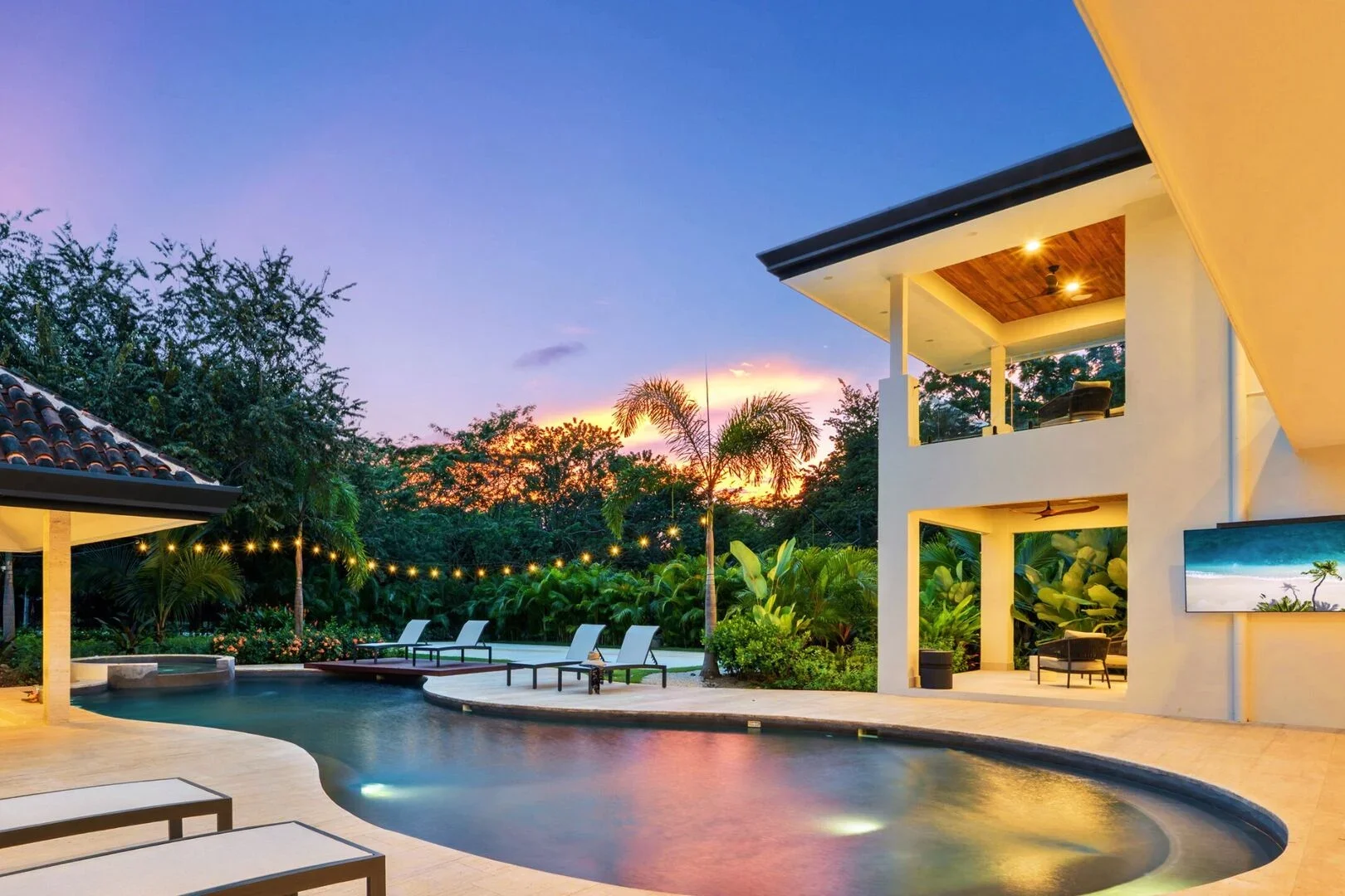 Luxury house with a swimming pool in a tropical setting during sunset, with trees, palm trees, outdoor seating, and string lights.