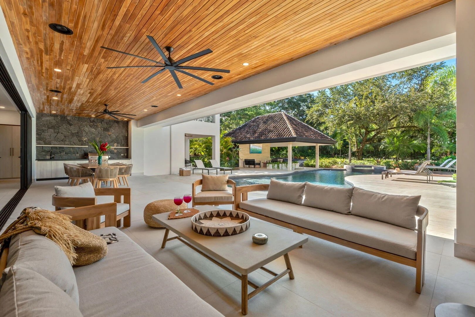 Open-air living space with a wooden ceiling, ceiling fans, and a view of a swimming pool surrounded by lush greenery, outdoor seating, and a small pavilion.
