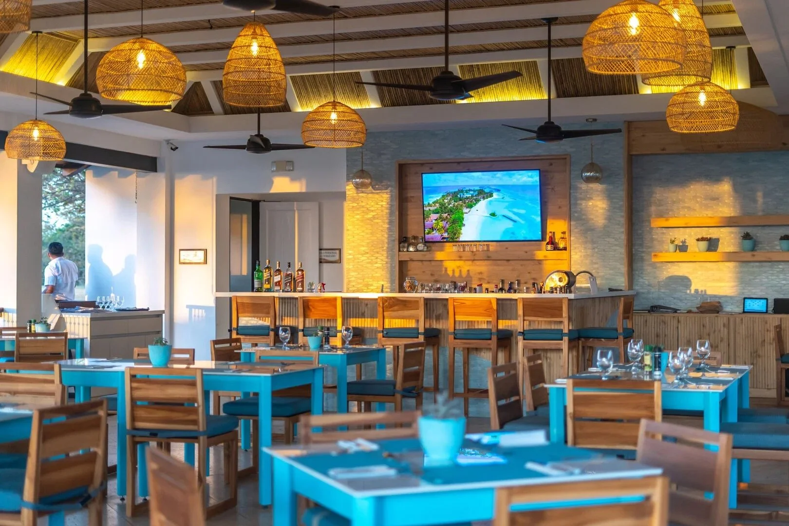 Interior of a beachside restaurant with wooden dining tables, some painted blue, set with glassware and napkins. There is a bar with stools and a large flat-screen TV showing an aerial view of a beach and ocean. Wicker pendant lights and ceiling fans